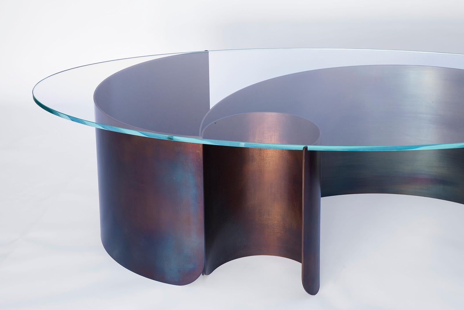 Rolled sheets of steel curl and peel away beneath a transparent oval glass surface, capturing the dynamic moment of a cresting wave. The vibrant multicolored steel surface is created through a unique heat treatment process through which molecules