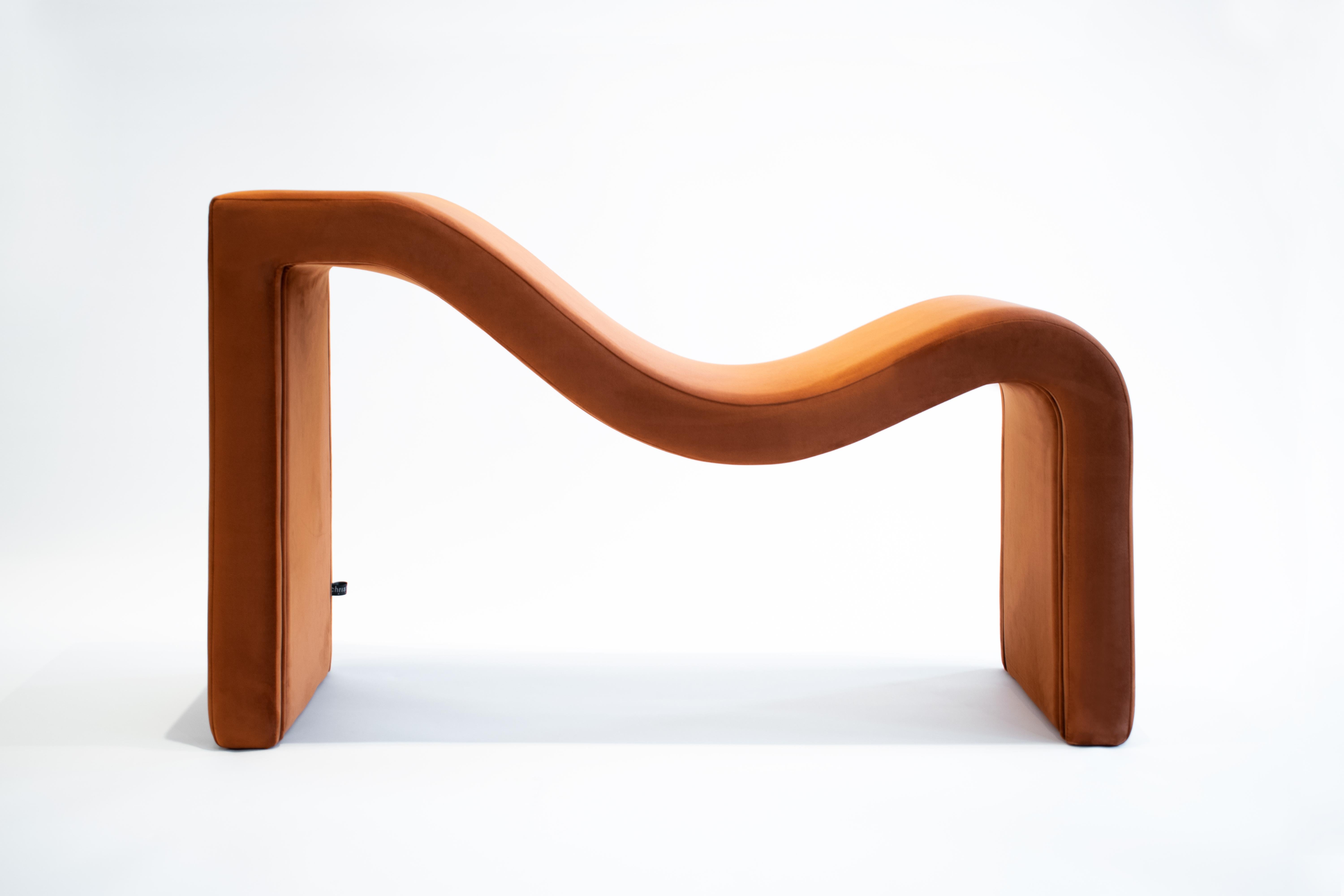 Revisiting the design of its predecessor the design of this single bench is inspired by the summer tides of the mediterranean during the sunset. Covered in macro plush fabric in a sun-kissed terracotta hue, the bench combines sharp and gently curved