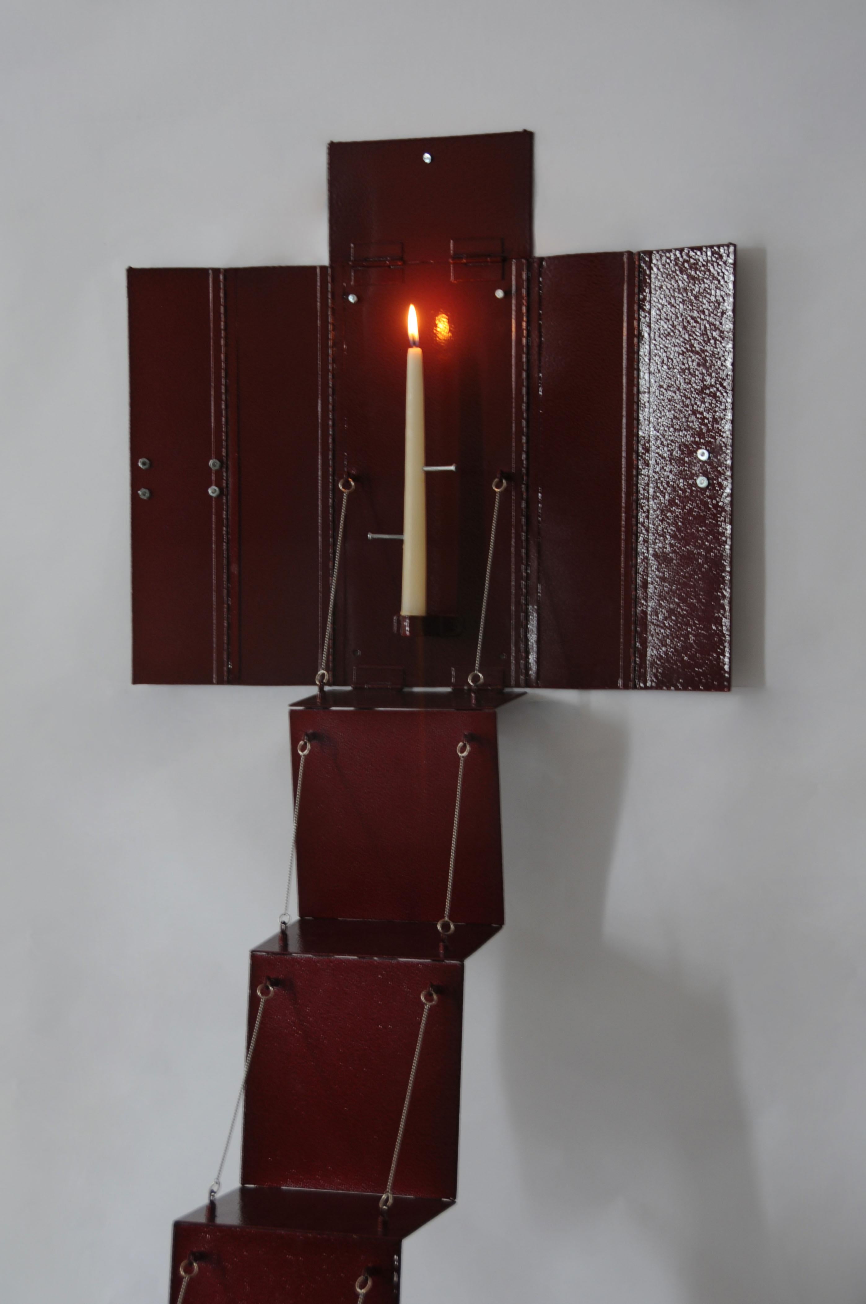 Ein Kerzenständer aus blutrotem Stahl ist als Altartafelbild an der Wand angebracht, das sich zusammenklappen lässt, um einen medizinischen Schrank zu simulieren. Die Kerze, die von der Metallstruktur getragen wird, wird von Nägeln in verschiedenen