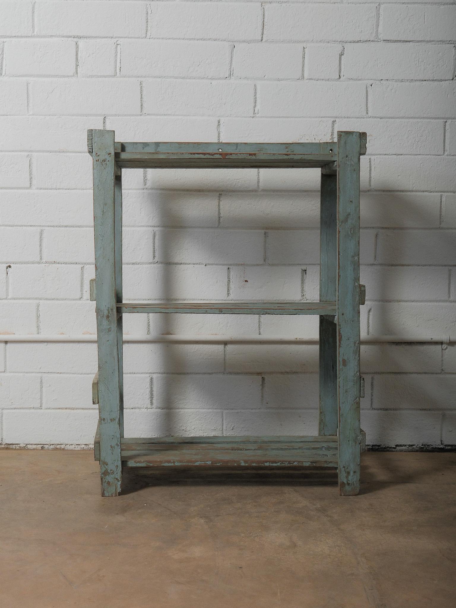 A quiet study in proportion and patina, this painted wooden shelf presents itself as a sculptural framework rather than a traditional storage piece. Its washed blue surface, worn through years of use, reveals layered tones and textures that lend the