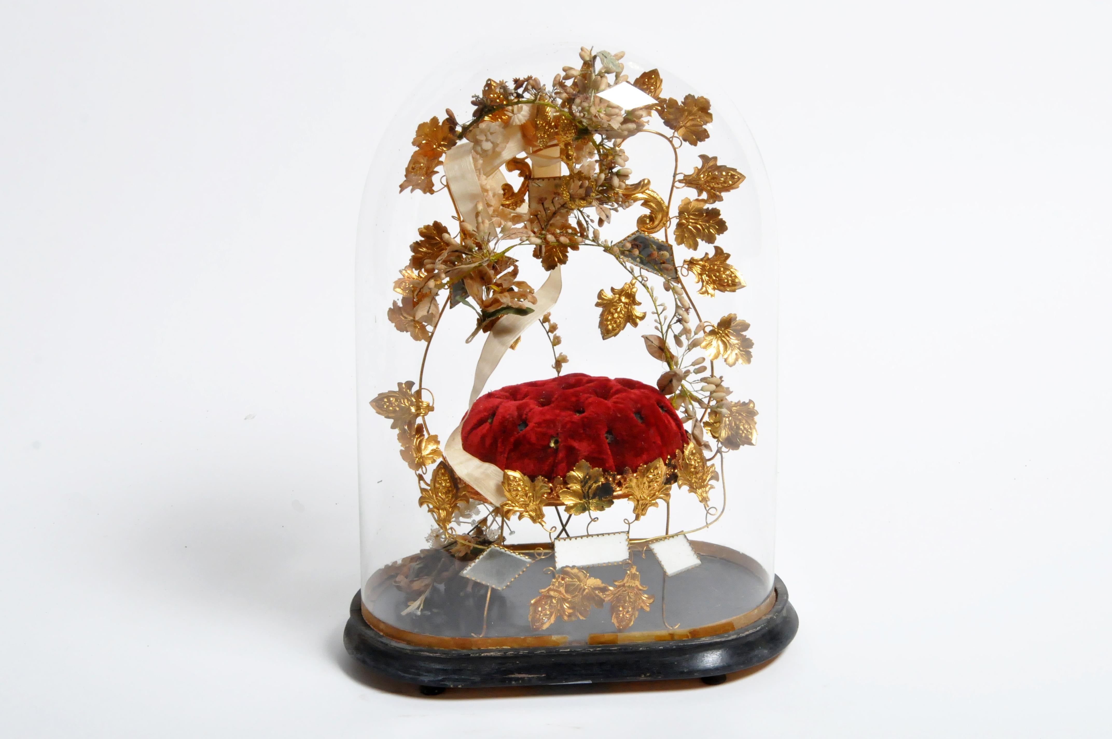 Quite the curiosity, this glass dome is more like a time capsule. Inside of the glass dome is an upholstered cushion with a frame and legs made of gilt foliage, porcelain flowers, and mirrored glass. A bird-form finial holding a wreath of laurel in