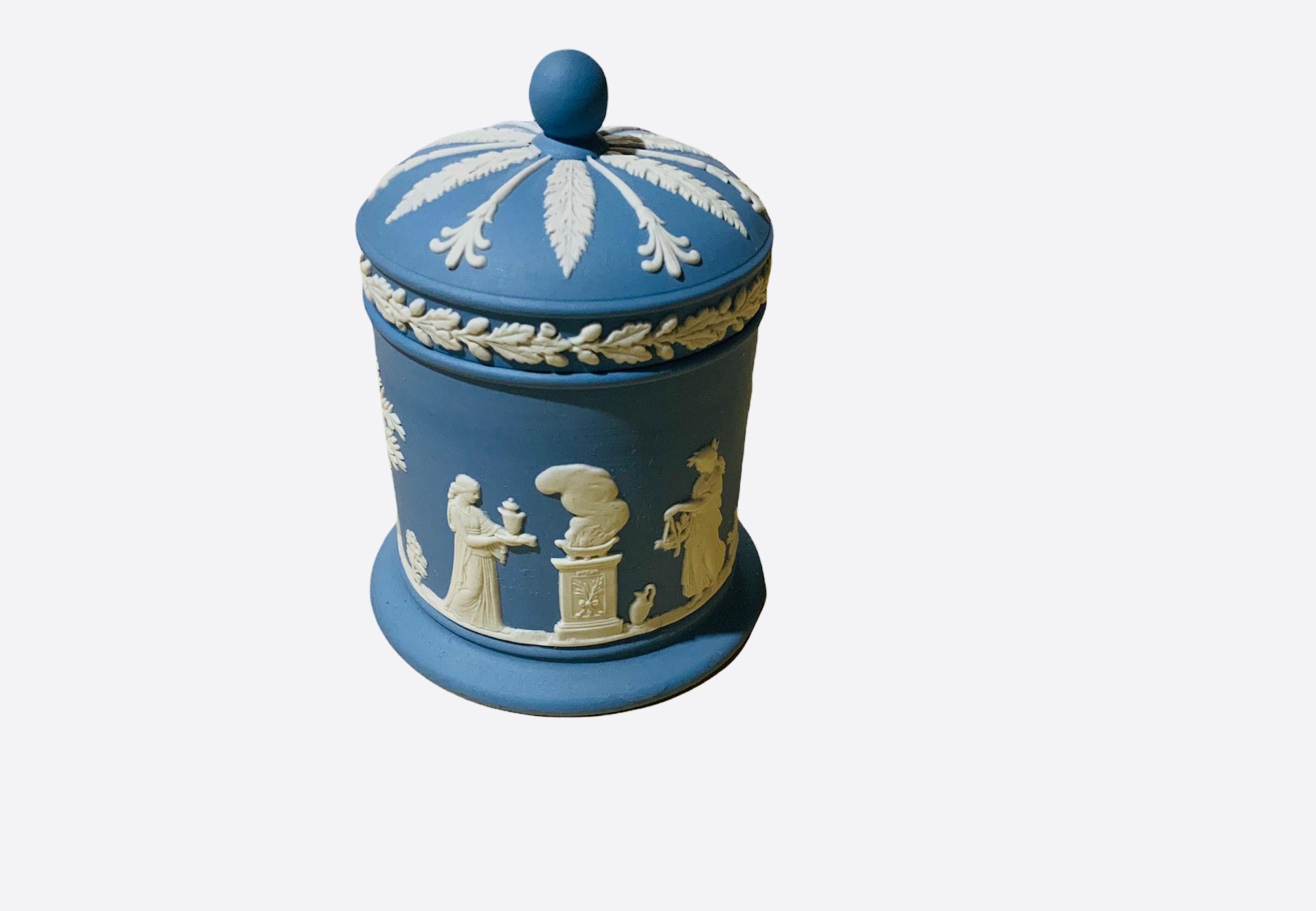 This is a Wedgewood Canister/Tobacco cylindrical lidded Jar with a blue background. It depicts a continuos Greek/Roman white relief scenes. In one scene, it is represented the goddess Artemis and Aphrodite between a winged cherub in a pedestal. The