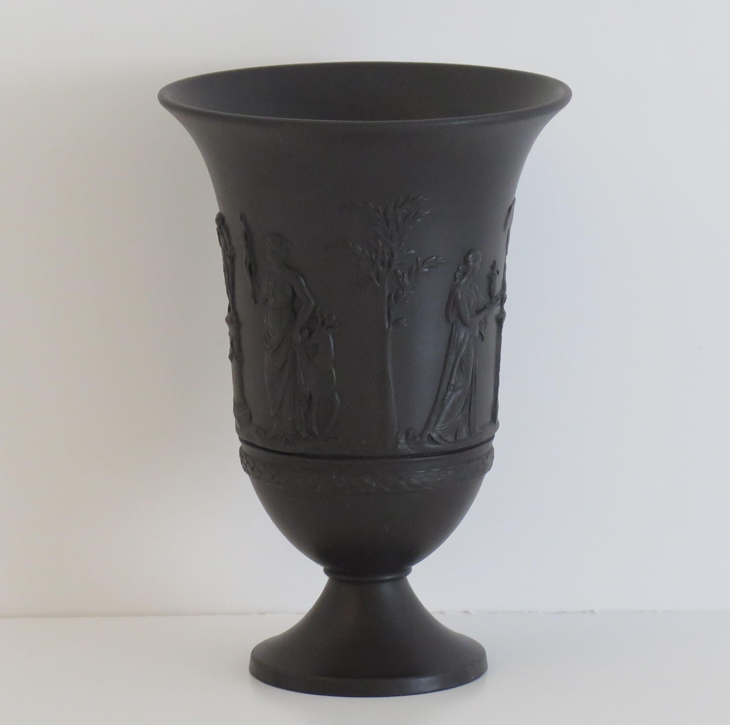 This is a good black basalt footed Vase, made by Wedgwood and dating to the early 20th century. 

The vase is well potted on a low foot with an everted rim.

The decoration shows classical figures, statues and trees in relief moulded form as