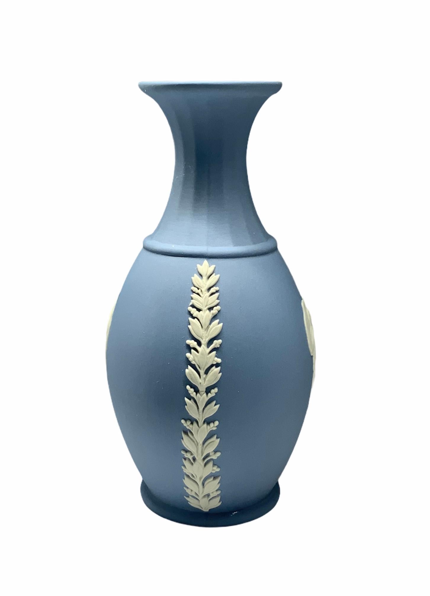 This is a Wedgwood blue porcelain vase with a trumpet fluted neck. It is adorned with two cameos of angel nymphs in the front and back of the bulbous body. The lateral sides are decorated with a vertical garland of laurel branches. Under the base is