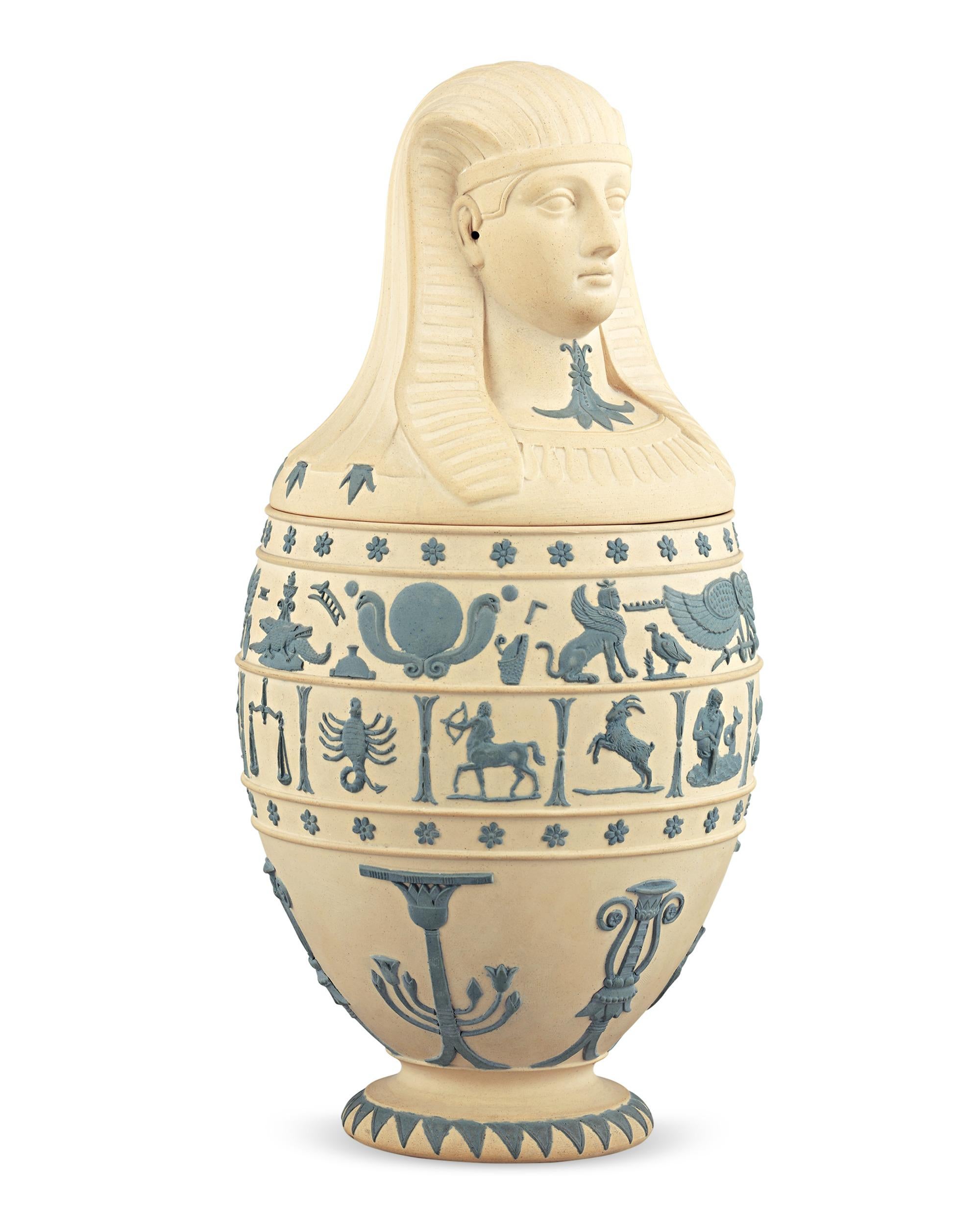 This 19th century Wedgwood caneware vessel takes the form of an Egyptian canopic jar, modeled after the important containers used for afterlife preparation during the process of mummification. Two central friezes of Egyptian artistic motifs and