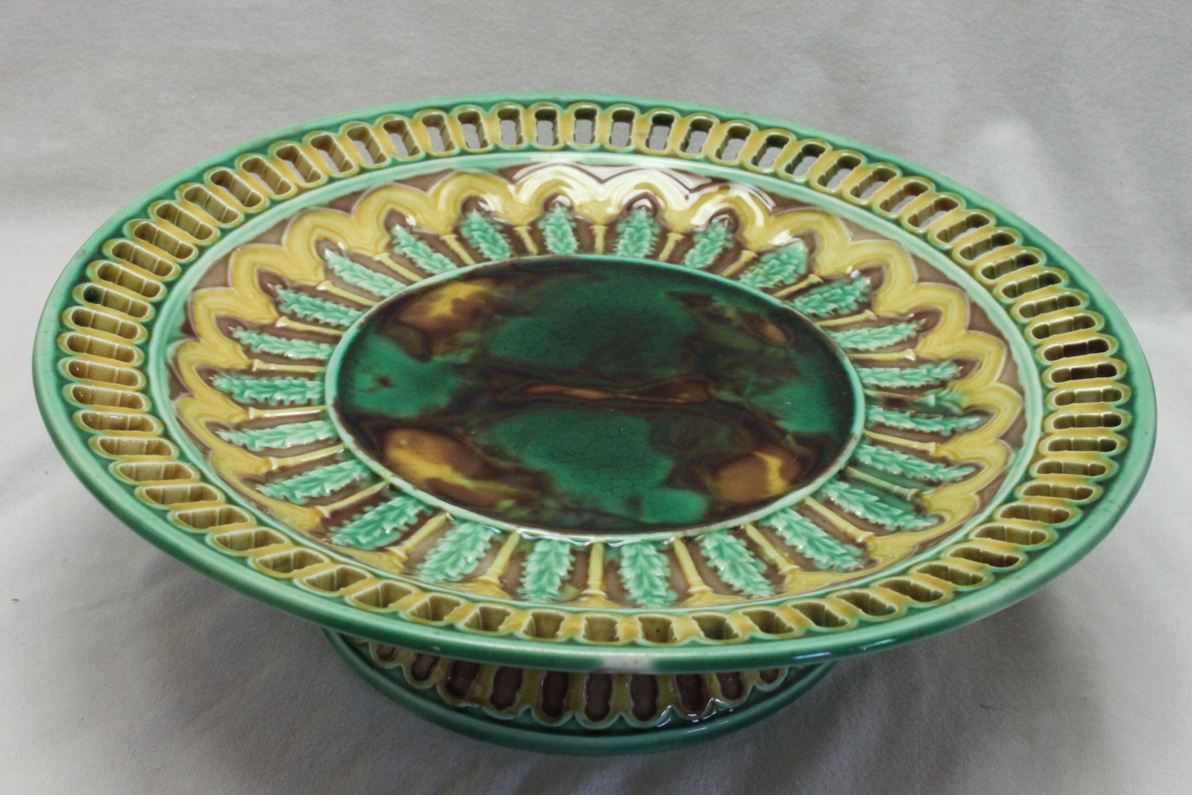 This Wedgwood majolica comport is decorated with green, brown and yellow majolica glazes around a central panel of mottled glazes. The reticulated rim is echoed in the blind moulding to the foot. The diameter is 210 mm (8.25 inches) and it stands 64