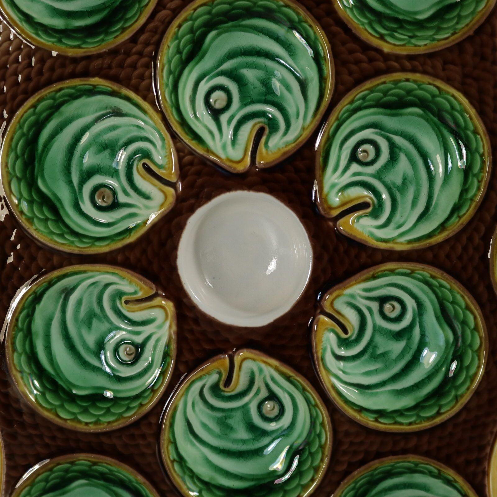 Wedgwood Majolica oyster plate which features twelve wells modelled as fish heads, surrounding a central white well. Brown ground molded as fish scales. Colouration: green, brown, yellow, are predominant. The piece bears maker's marks for the