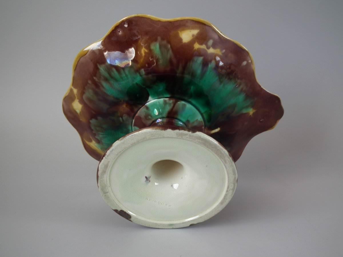 Compota de mayólica Wedgwood con hojas de vid y uvas sobre un fondo de tejido de cestería. Coloración: predominan el verde, el marrón y el azul. La pieza lleva marcas de fabricante de la cerámica Wedgwood.