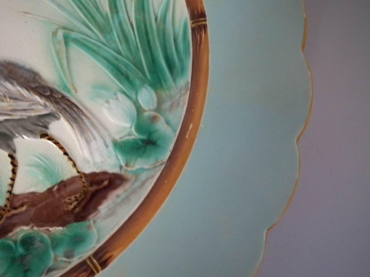 Wedgwood Majolica plate which features a heron at the edge of a pond. Designed by Joseph Theodore Deck. Colouration: pale blue, green, cream, are predominant. The piece bears maker's marks for the Wedgwood pottery. Bears a pattern number, '864'.