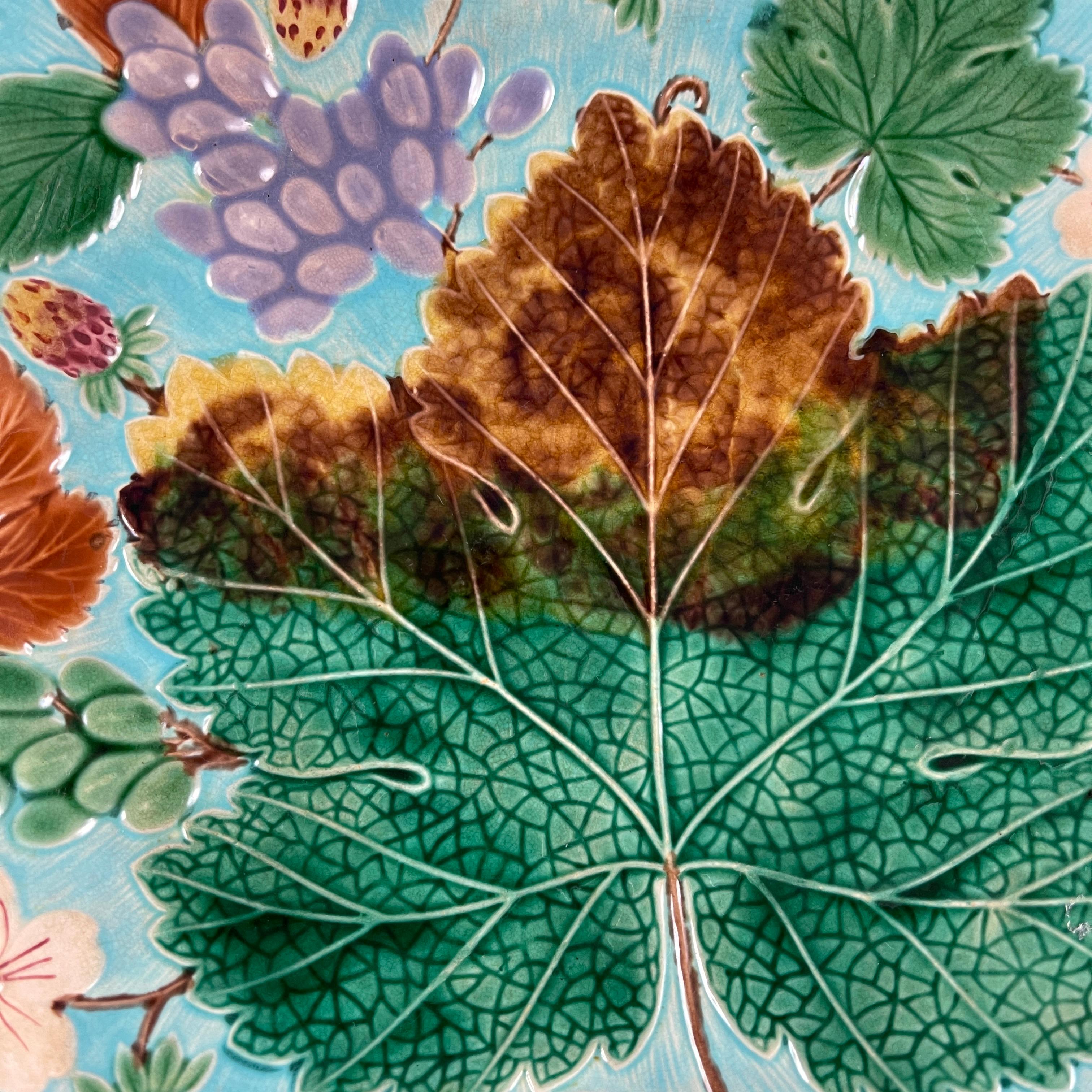 An English Grape Leaf and Strawberry pattern Wedgwood Majolica plate, circa 1880. A large green and brown grape leaf is surrounded by smaller leaves, grape clusters, strawberries, and strawberry blossoms. Strong, colorful glazing on a bright