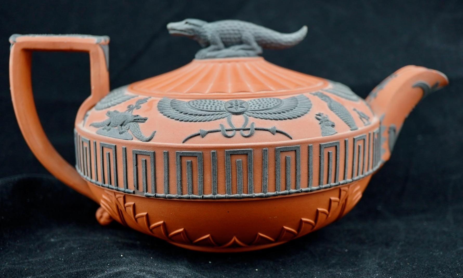 Wedgwood Rosso Antico Egyptian Revival Teapot. The teapot surmounted by figural black crocodile finial, radiating ribbed lid, body decorated with Egyptian motifs including winged sphinxes, crocodiles, burial jars, animals, keyed border to waist,