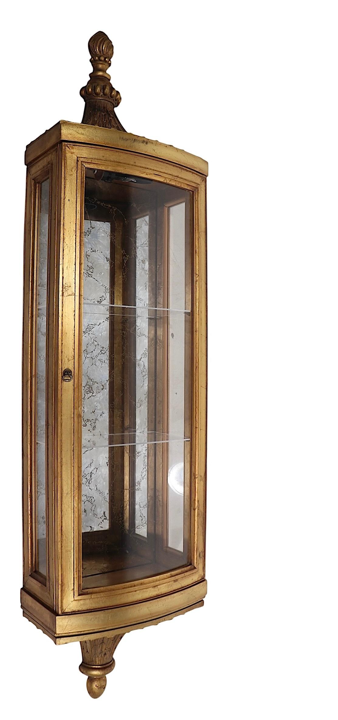 Glamorous Hollywood Regency style wall hanging display curio cabinet, vitrine, showcase, executed in carved wood in gold pilot finish, with a curved glass door, and a marbleized mirrored interior. The case comes with two lucite shelves, possibly