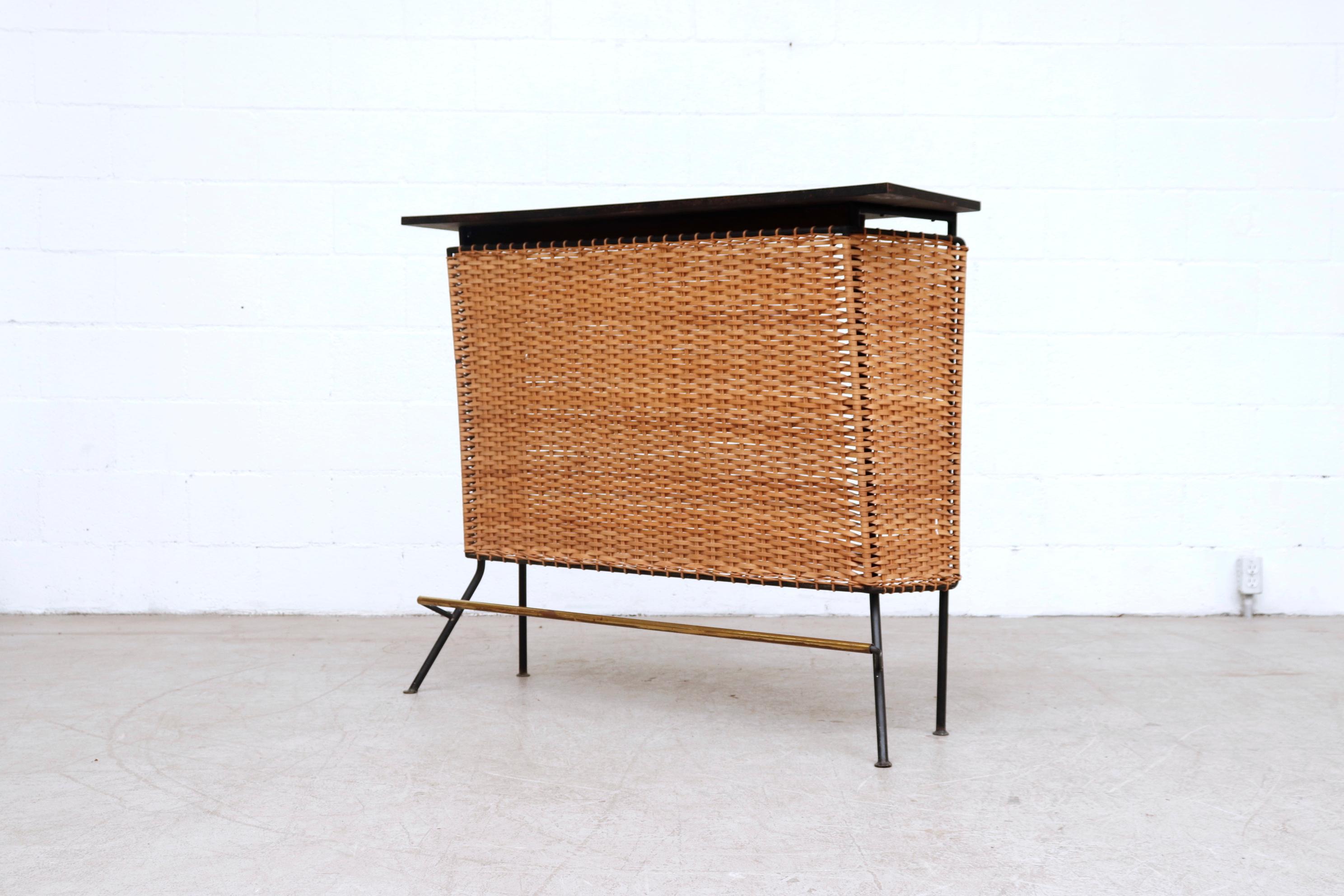 Incredible midcentury Frederick Weinberg style rattan bar with black enameled metal frame, rattan body and brass footrest. In original condition with visible wear including some rattan breakage, and visible patina to the metal. Wear is consistent