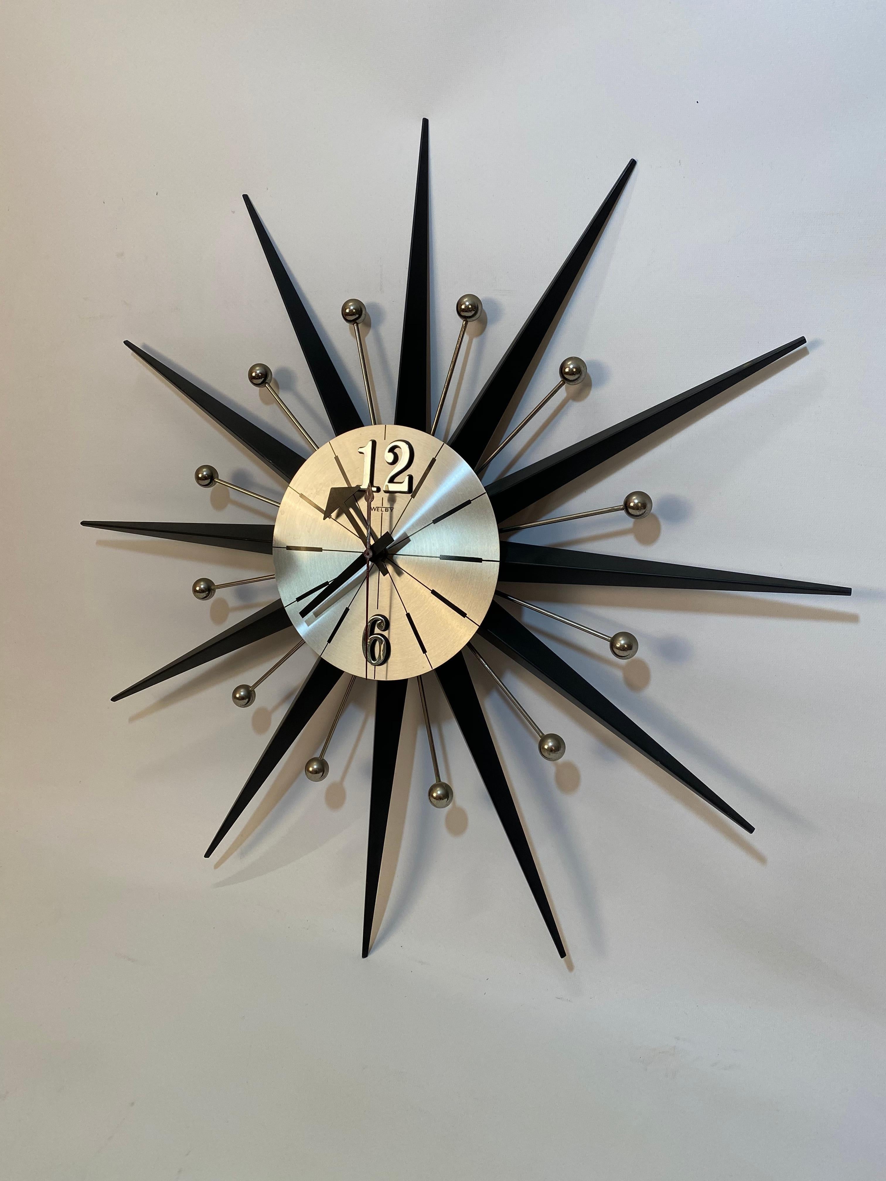 Welby sunburst wall clock. Black radiating arms with black numbers hour and minute hands on a brushed aluminum clock face, circa 1960. Battery powered (Battery C). Good working condition. No visible losses, scratches. Black arms are