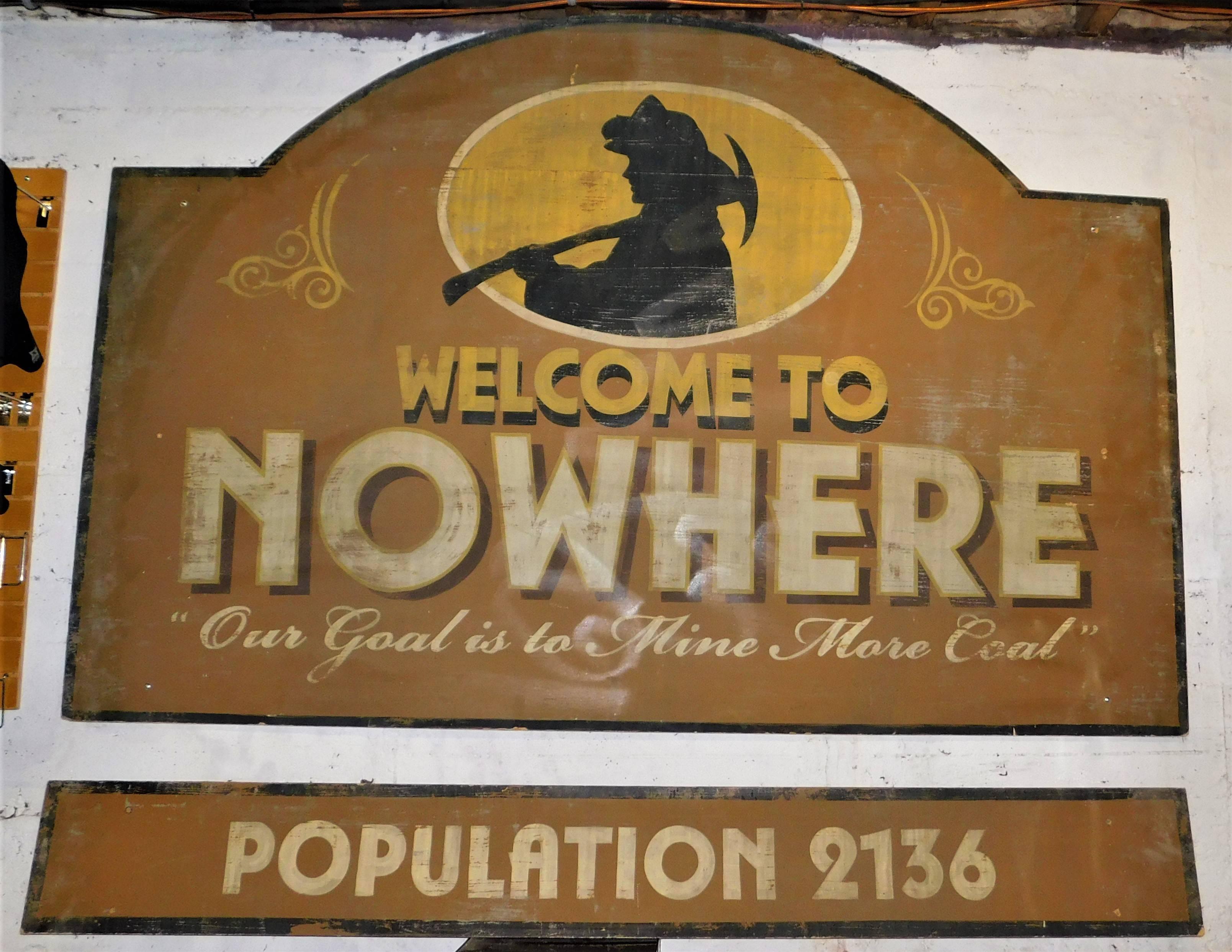 Large wooden town sign Welcome to Nowhere, has a coal miner on it holding a pick ax wearing a helmet. 