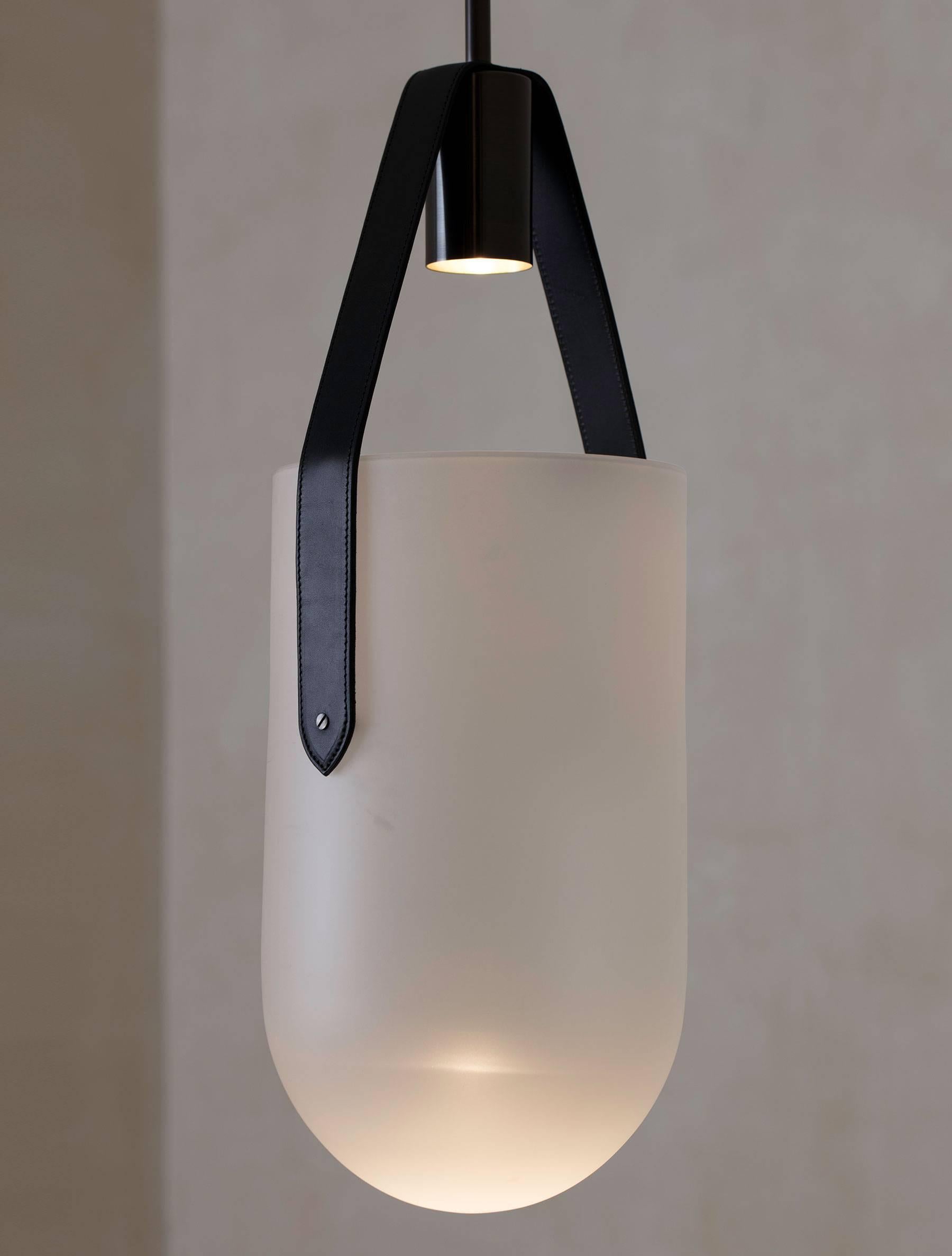 A spot bulb shoots light into a sandblasted glass dome that is suspended by a leather strap and brass hardware. The fixture creates a warm glow inside the glass as well as casting dramatic shadows onto the ceiling. UL Certified.