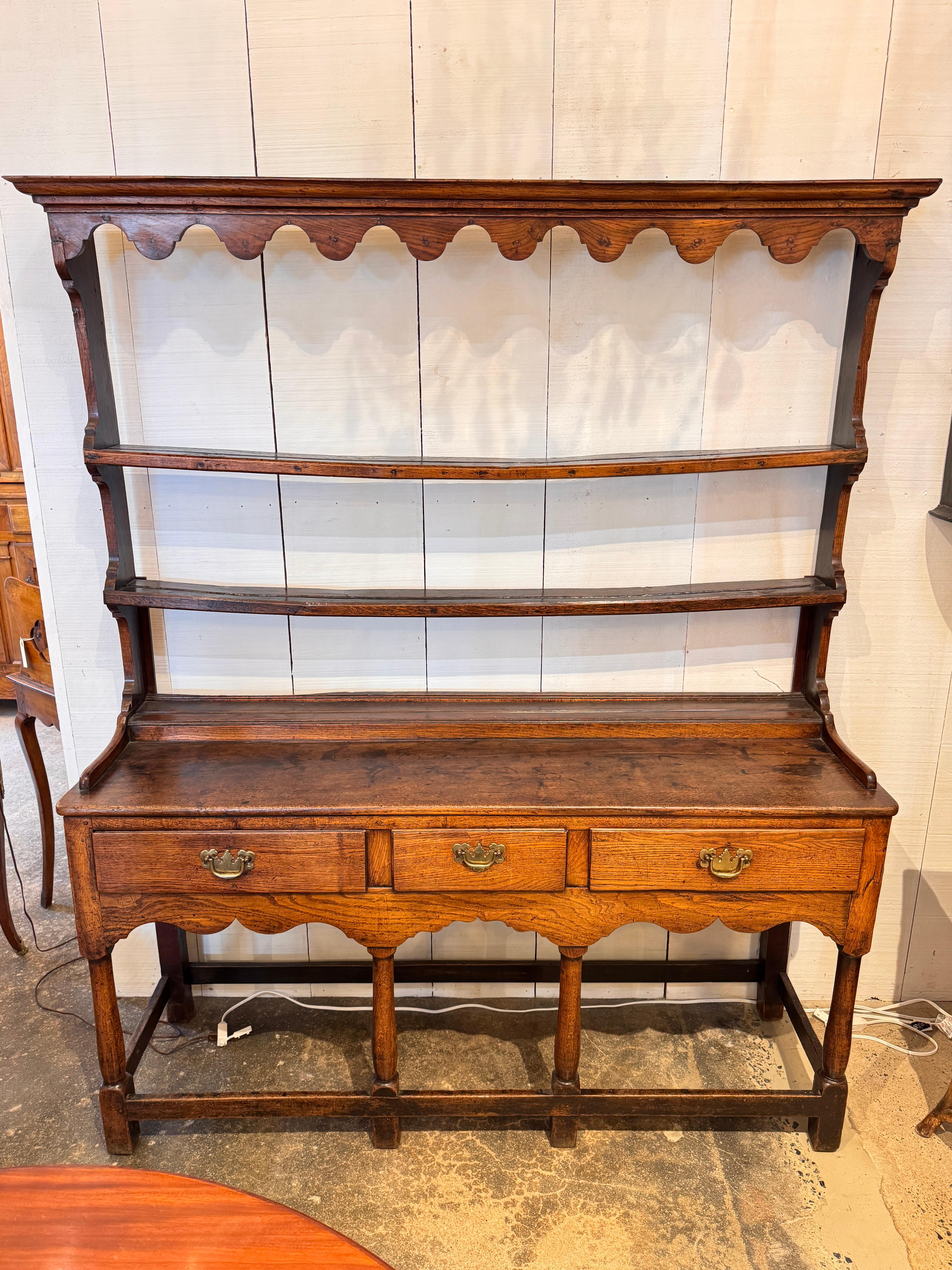 Welsh oak dresser featuring a molded cornice over a scalloped plate rack with two open shelves. The base is fitted with three drawers above a shaped apron, raised on block legs joined by stretchers. The rich oak surface shows a warm patina.
