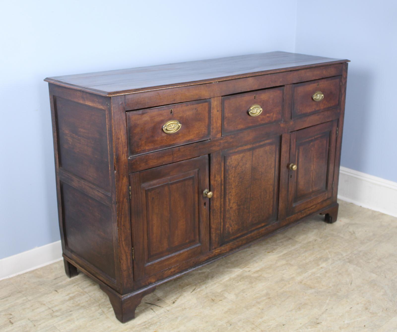 A handsome and quite early dresser base, buffet, or cupboard in dark oak with mahogany crossbanding. The center section is closed with a front panel and the interior is two equal storage sections with a half shelf. Doors shut well with turn latches