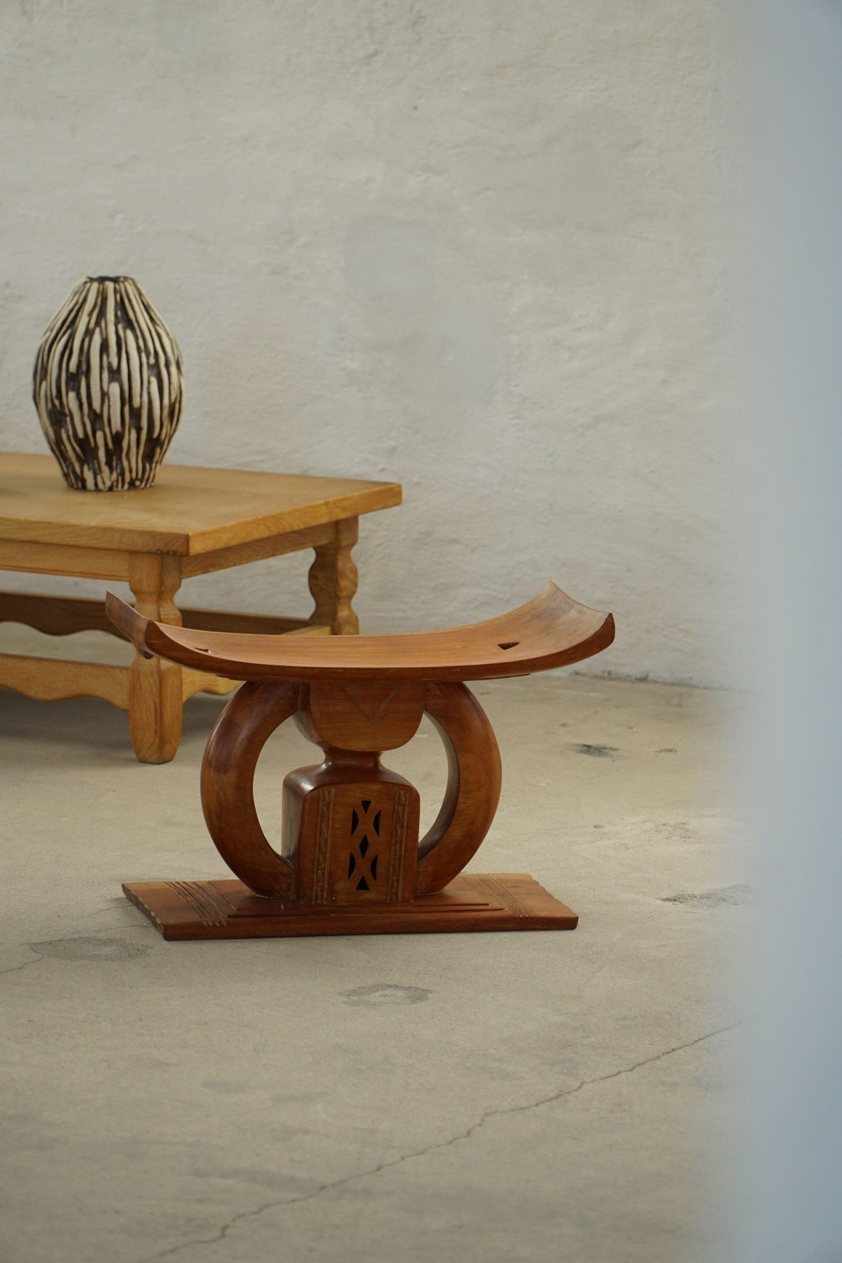 This sculptural stool is carved from a single block of solid wood, showcasing a distinctive, monolithic form. The wide, curved seat rests on a central arched support flanked by two end blocks — creating a grounded, rhythmic silhouette with both