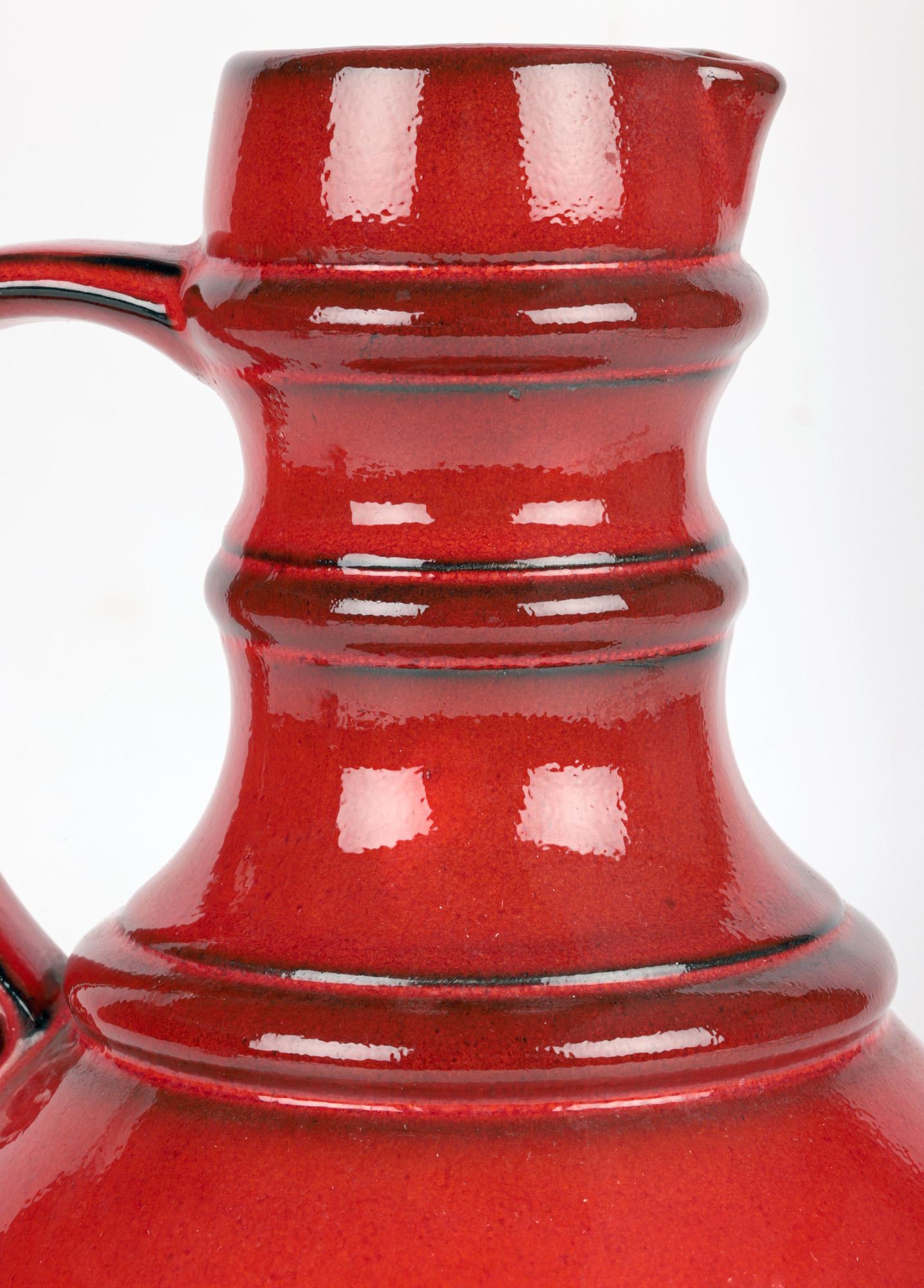 An exceptional West-German mid-century art pottery red lava glazed pitcher vase dating from around 1950-60. The ceramic pitcher stands raised on a narrow round foot with a round bulbous body and shaped tall narrow three ring neck with a short funnel