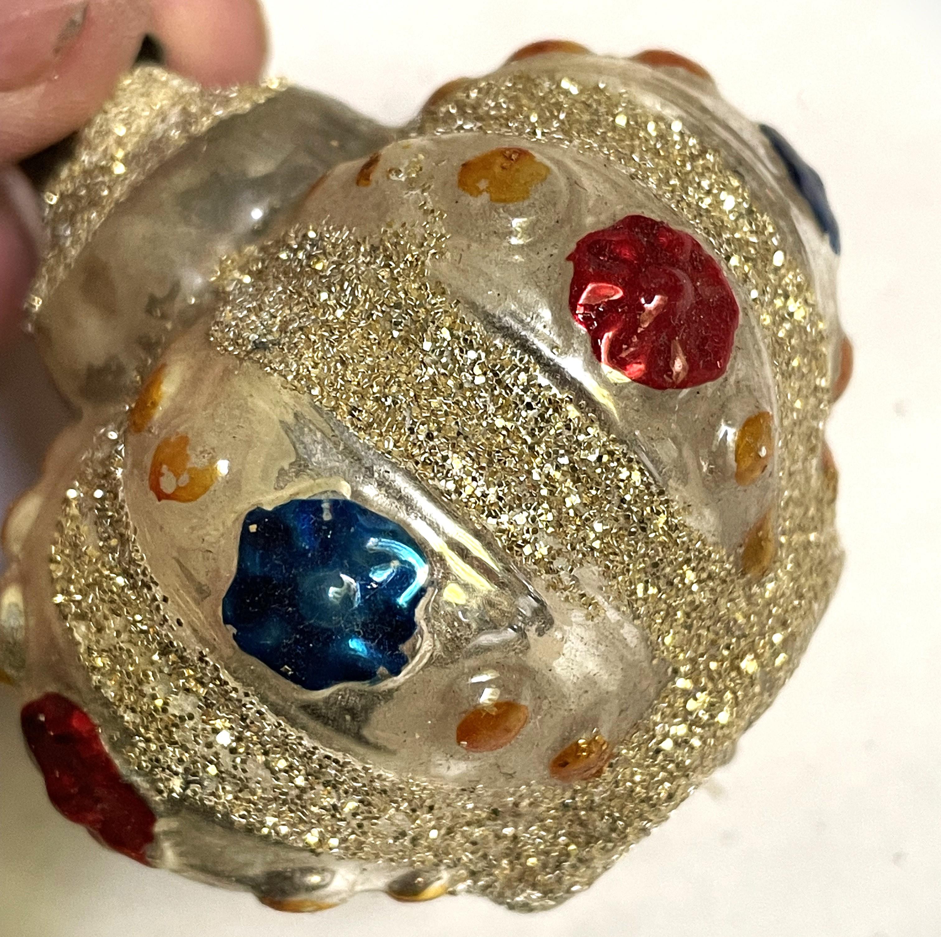 Voici une jolie décoration à l'allure très royale, avec une couronne en 3D composée de crêtes bulbeuses. Chaque crête est ornée d'un gros point rouge et bleu alterné avec de petits points dorés au-dessus et en dessous.

Cet ornement est en bon état,