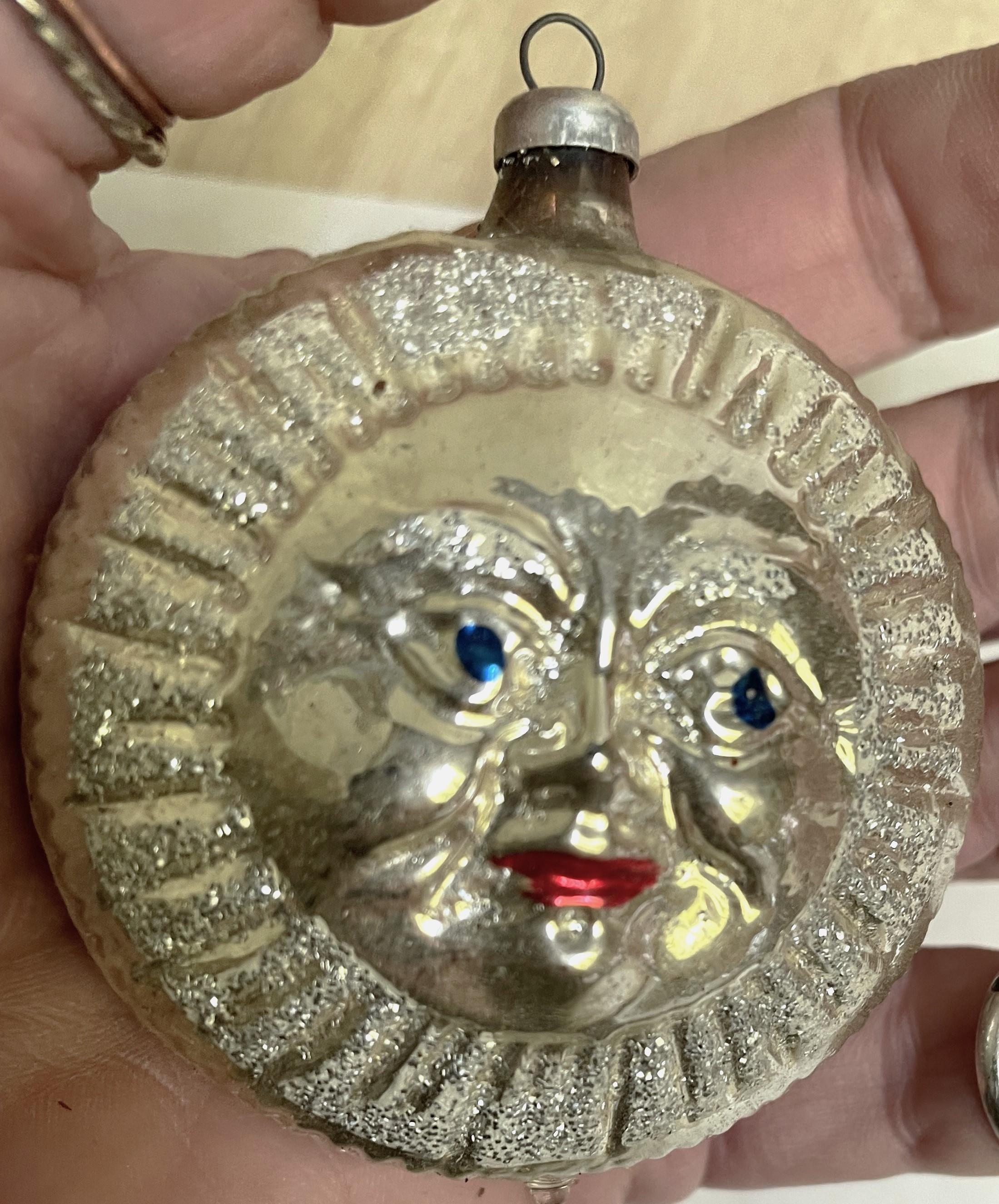 Si tratta di un intrigante ornamento in vetro al mercurio argentato con occhi blu e labbra rosse, circondato da brillantini bianchi e lucenti, che sembra una luna che fa capolino da un cielo pieno di neve. Funzionerebbe benissimo come decorazione