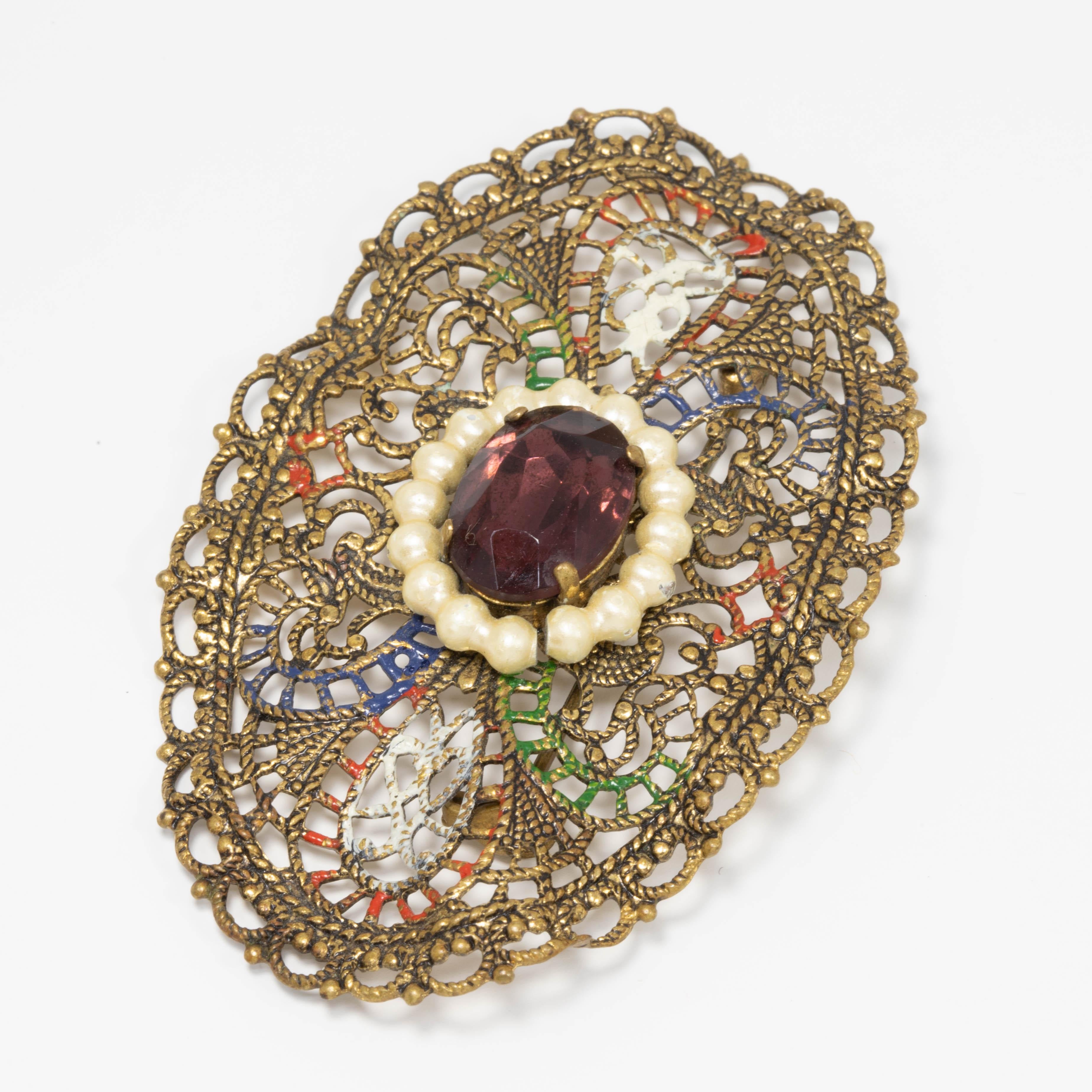 A stylish vintage German pin. Features a filigree brasstone metal setting with an amethyst crystal centerpiece, accented with faux pearls around. The setting is painted with white, orange, blue, and green enamel.

Vintage, circa mid 1900s. Made in