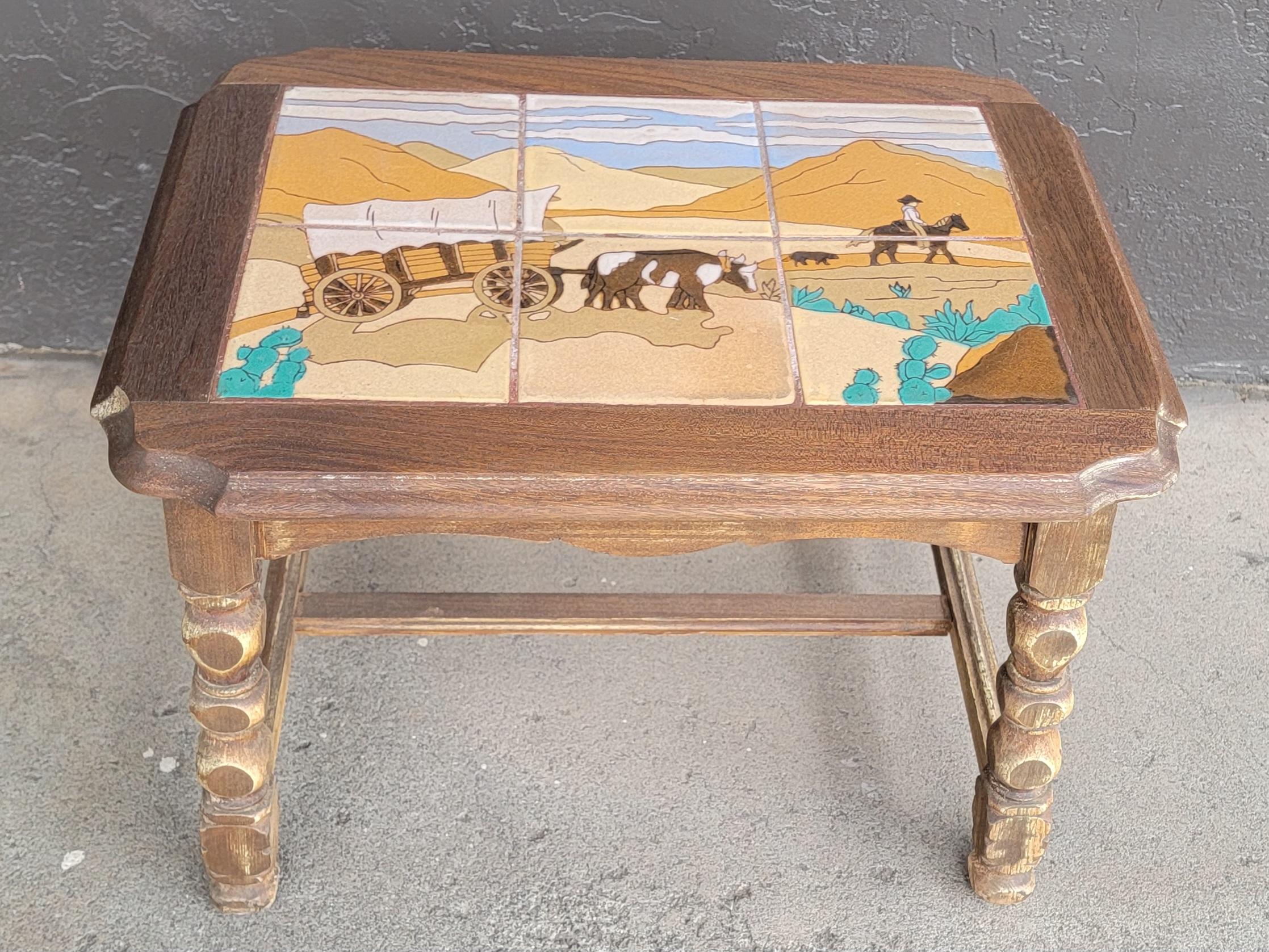 Western motif tile top table by Taylor Tile, California. Circa. 1930's. Depicts a covered wagon pulled by oxen in high desert country, a cowboy on horse with a dog. Cactus and mountain background. Six hand painted glazed tiles mounted to original