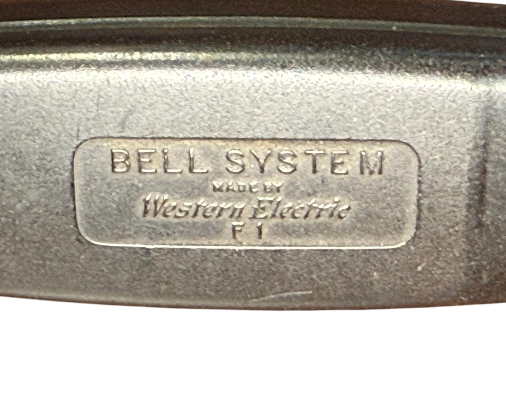Il s'agit d'un téléphone Western Electric Bell System Spacesaver. Elle représente un téléphone rotatif de couleur noire, modèle 211, avec son combiné F 1 et ses cordons. Le téléphone peut être fixé au mur. Elle est poinçonnée au dos, Bell System by