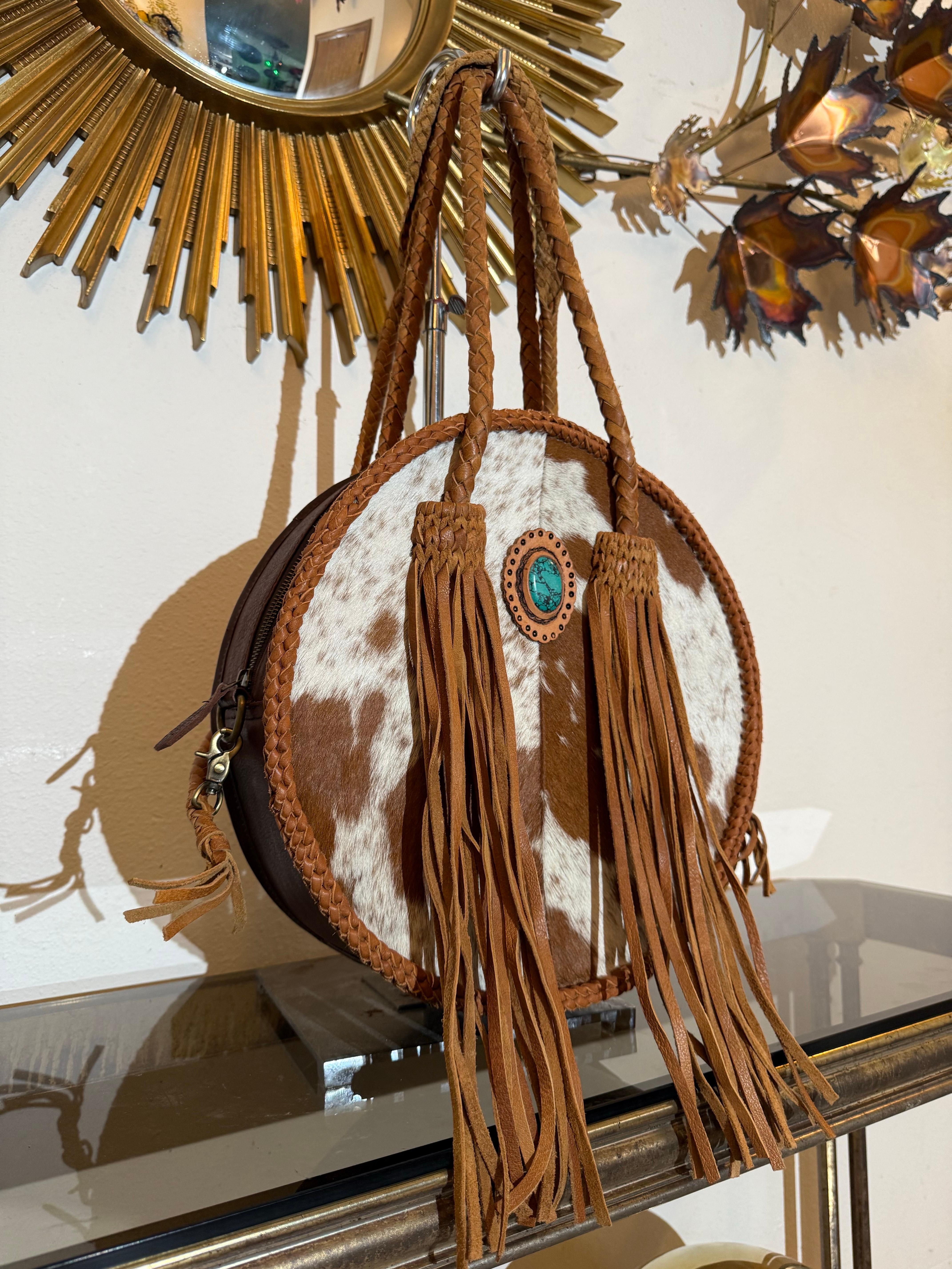 Western-Styled Cowhide Drum Handbag w Braid Handles and Leather Tassels:  A chic, rugged shoulder bag with whip braid style handles long enough to use as a shoulder bag. Faux turquoise decorative concho medallion. Brown and white patterned cowhide.