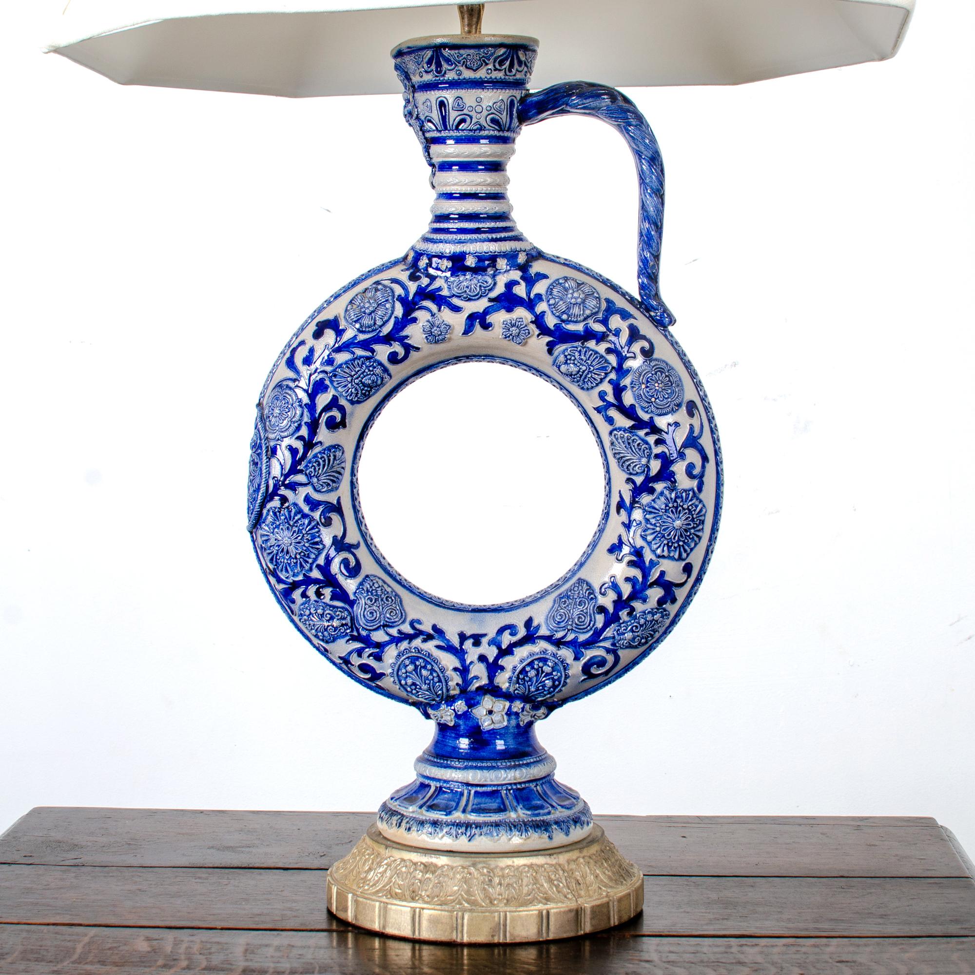 A late 19th or early 20th century Westerwald ring jug converted to a lamp.  Bellarmine face on tapering neck, with rope-twist handle, molded flowering vines and armorial roundel, with reeded foot.  Blue under salt glaze.

Jug measures 11 inches wide