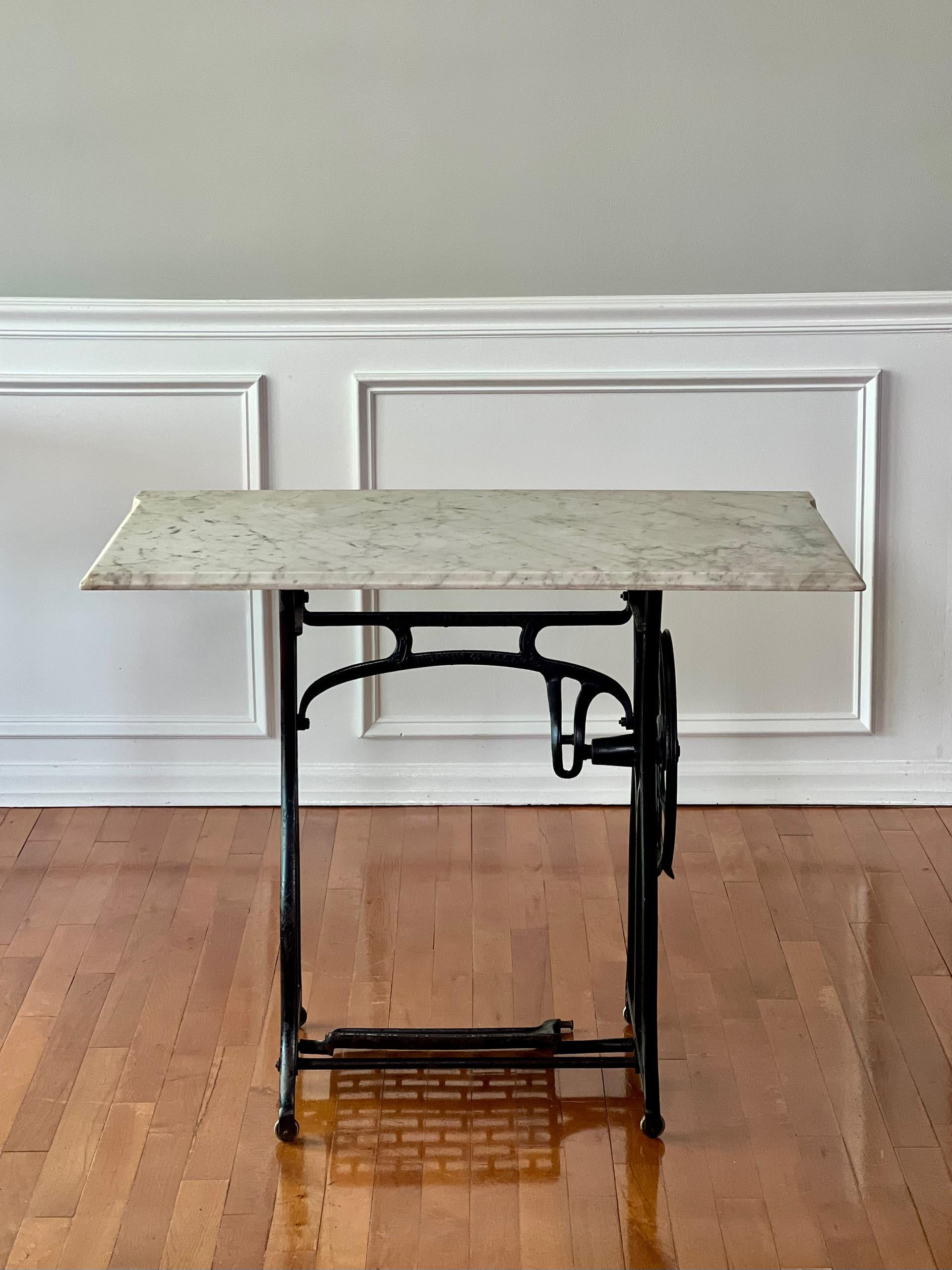 Early 20th century iron sewing machine table base with white marble top by Wheeler & Wilson of Bridgeport, CT. The rectangular marble top has gray veining and a beveled edge on three sides. The base has fixed position casters and is missing the