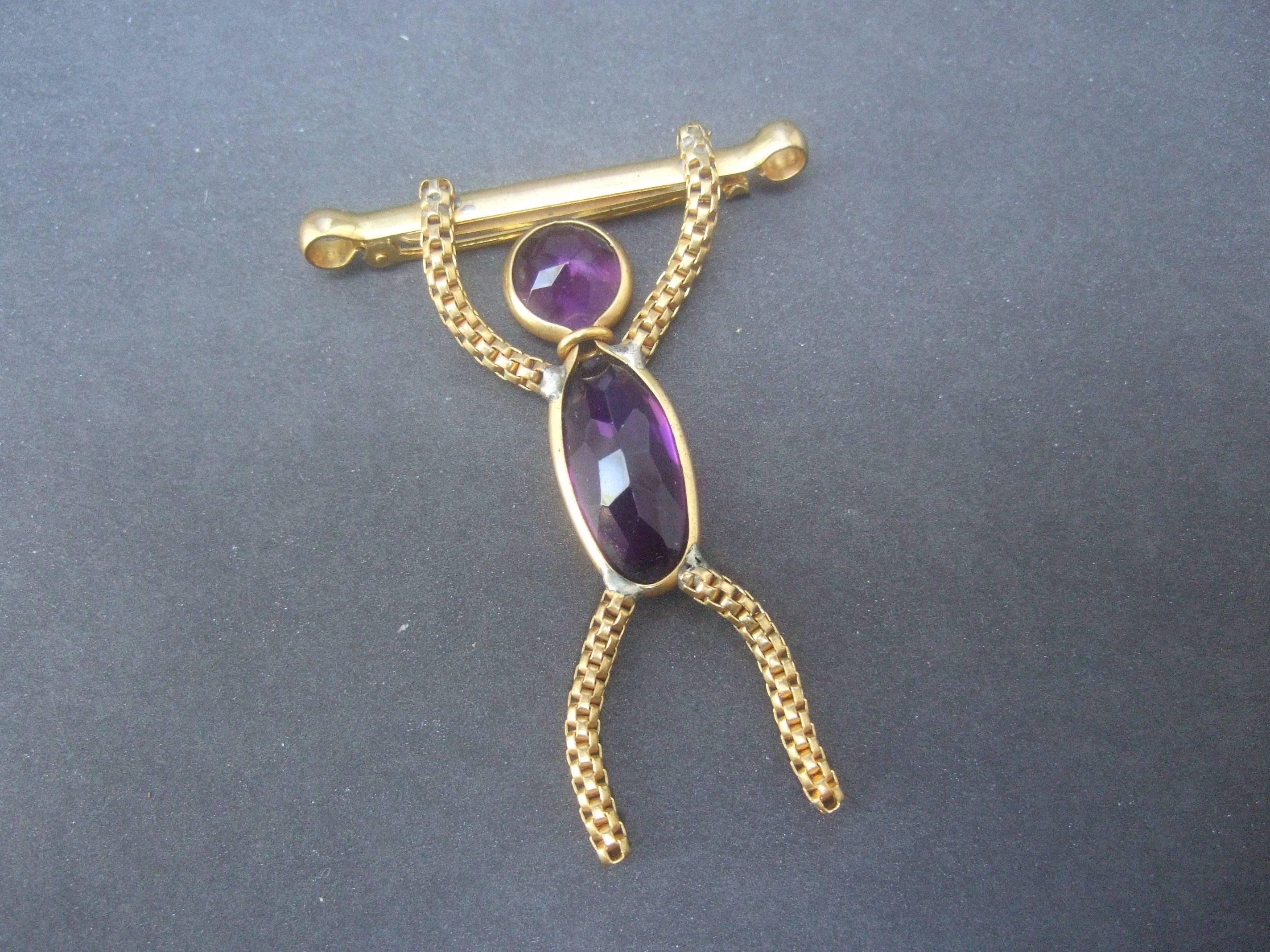 Whimsical figural hang man crystal brooch c 1960
The unique figural brooch is designed with an
andrognous figure that may be a man or monkey
figure suspended from a gilt metal bar 

The figure is embellished with an elongated 
amethyst color crystal