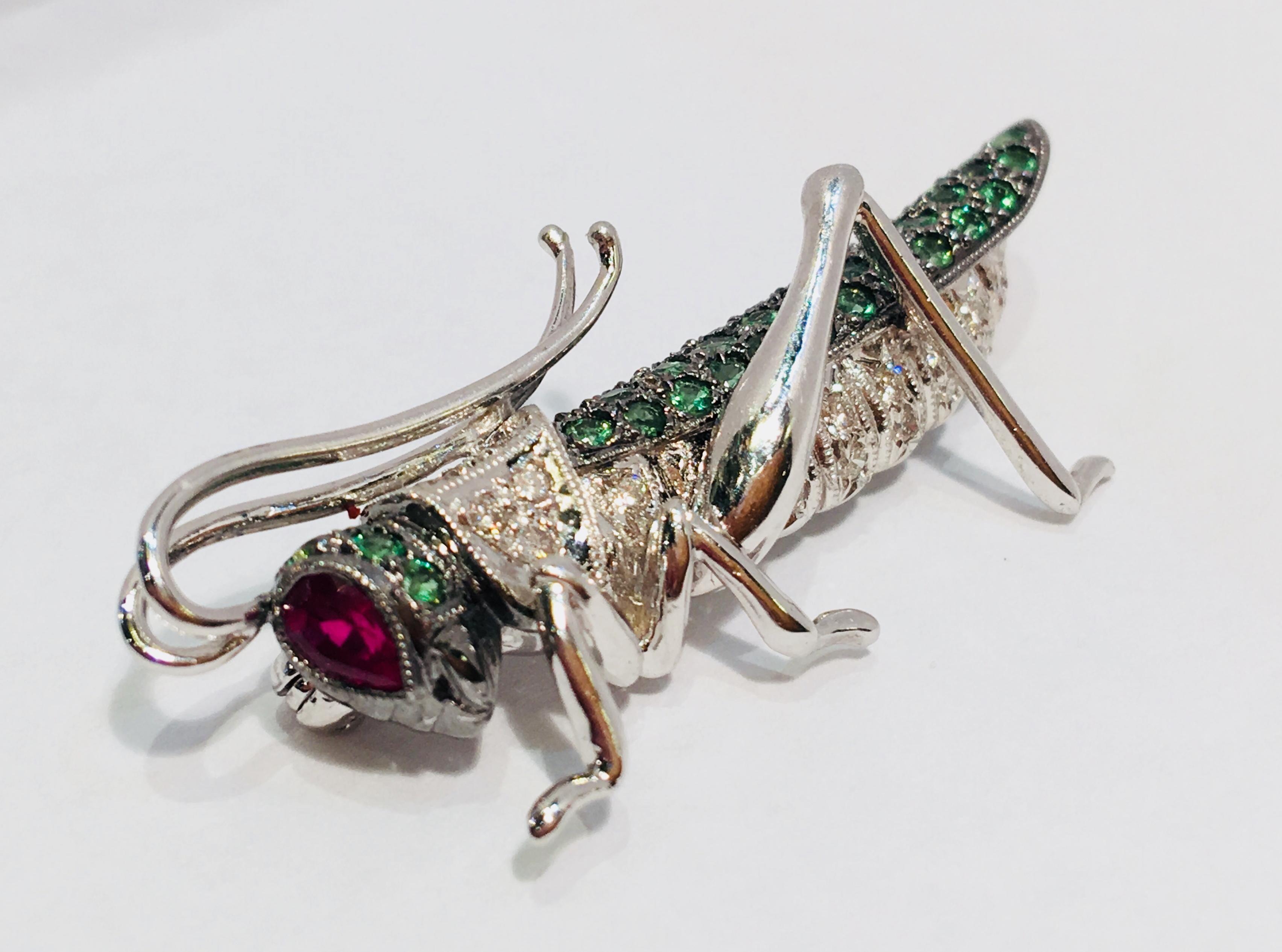 Adorable 18 karat white gold brooch pin in the shape of a whimsical grasshopper or cricket features a pave set round brilliant diamond body, pave set green tsavorite head and wing, and a pear shaped, bezel set ruby eye, as well as highly polished