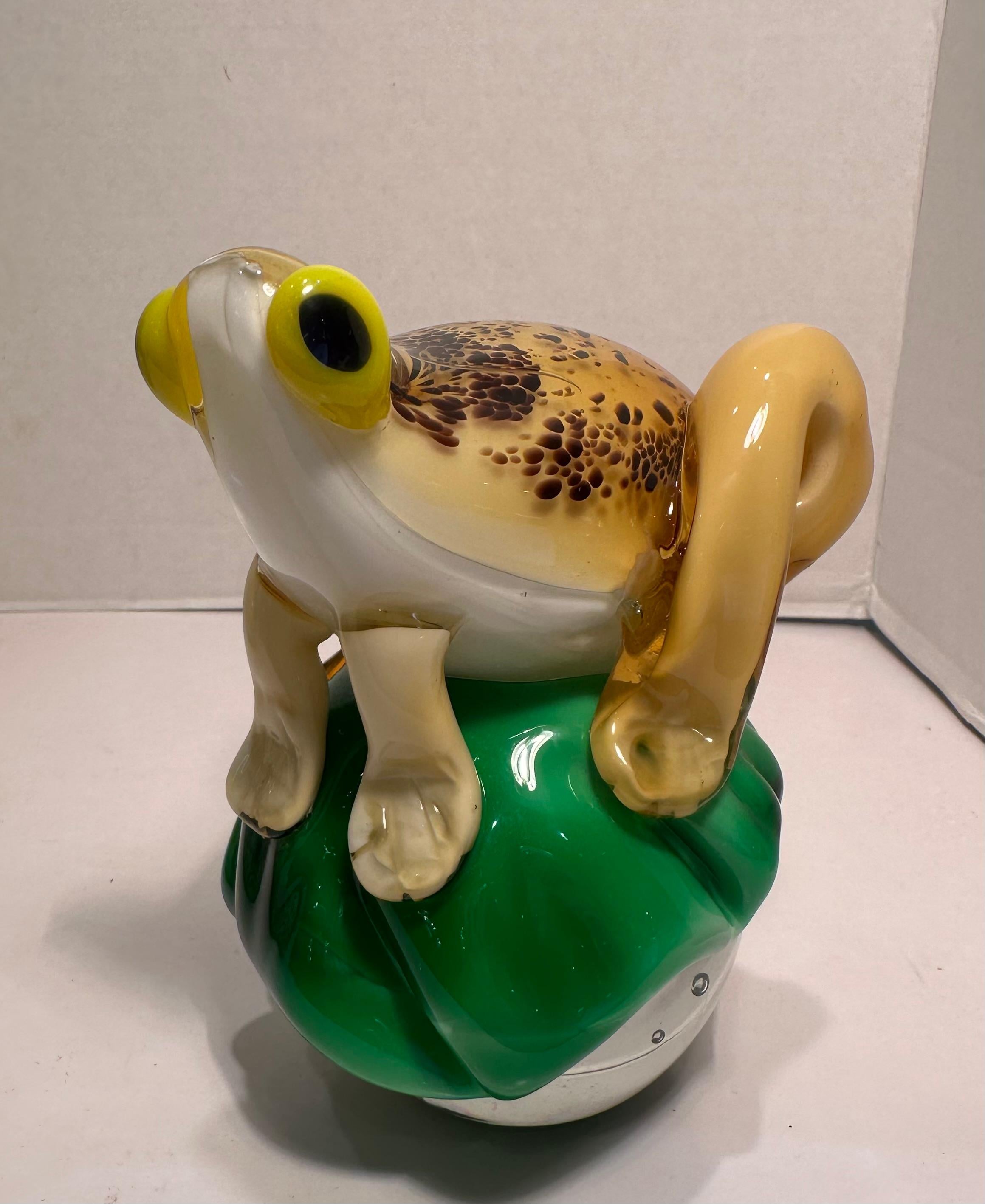 Sehr künstlerisch, lustig und skurril, handgefertigt und mundgeblasen in Italien, goldgelb mit braun gesprenkeltem Rücken, weißbäuchiger Kunstglasfrosch auf einem grünen Lilienpad, flankiert von einer bauchigen, blasigen Kunstglaskugel oder einem