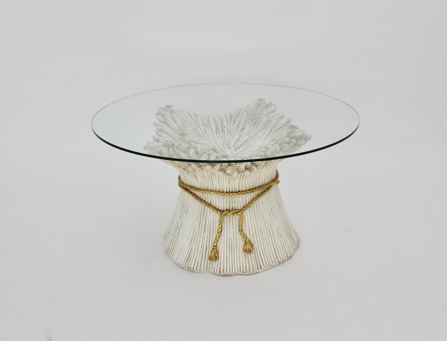 A white and gold Hollywood Regency Sheaf of Wheat coffee table, which was designed 1970s Italy.
The white and gold colored coated ceramic coffee table shows a sheaf of wheat bundled with a golden rope.
It features a clear glass top without