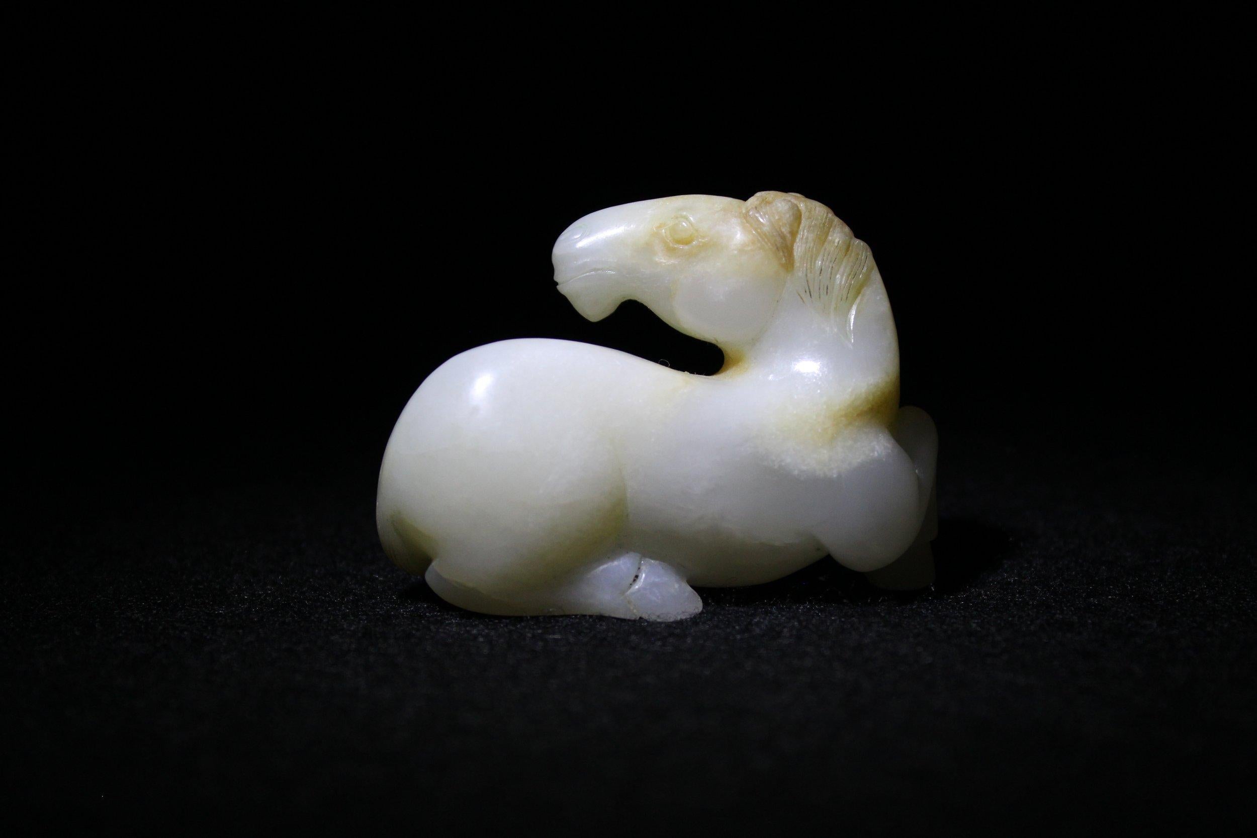 A resting horse has been carved from white jade with patches of russet. The long muscular body rests on four bent legs, and the head is turned around. 

The mane is embellished with long striated locks extending down the back of the neck, and the