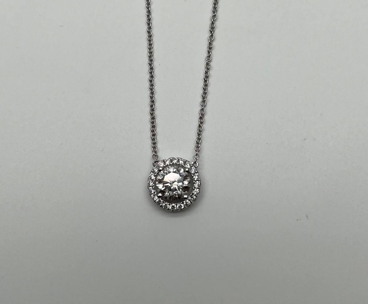 Cette pièce exquise présente un diamant rond central entouré d'un halo de diamants plus petits.

Le pendentif est serti dans une élégante monture en or blanc, rehaussant l'éclat des pierres.

La chaîne est délicate mais robuste, complétant le design