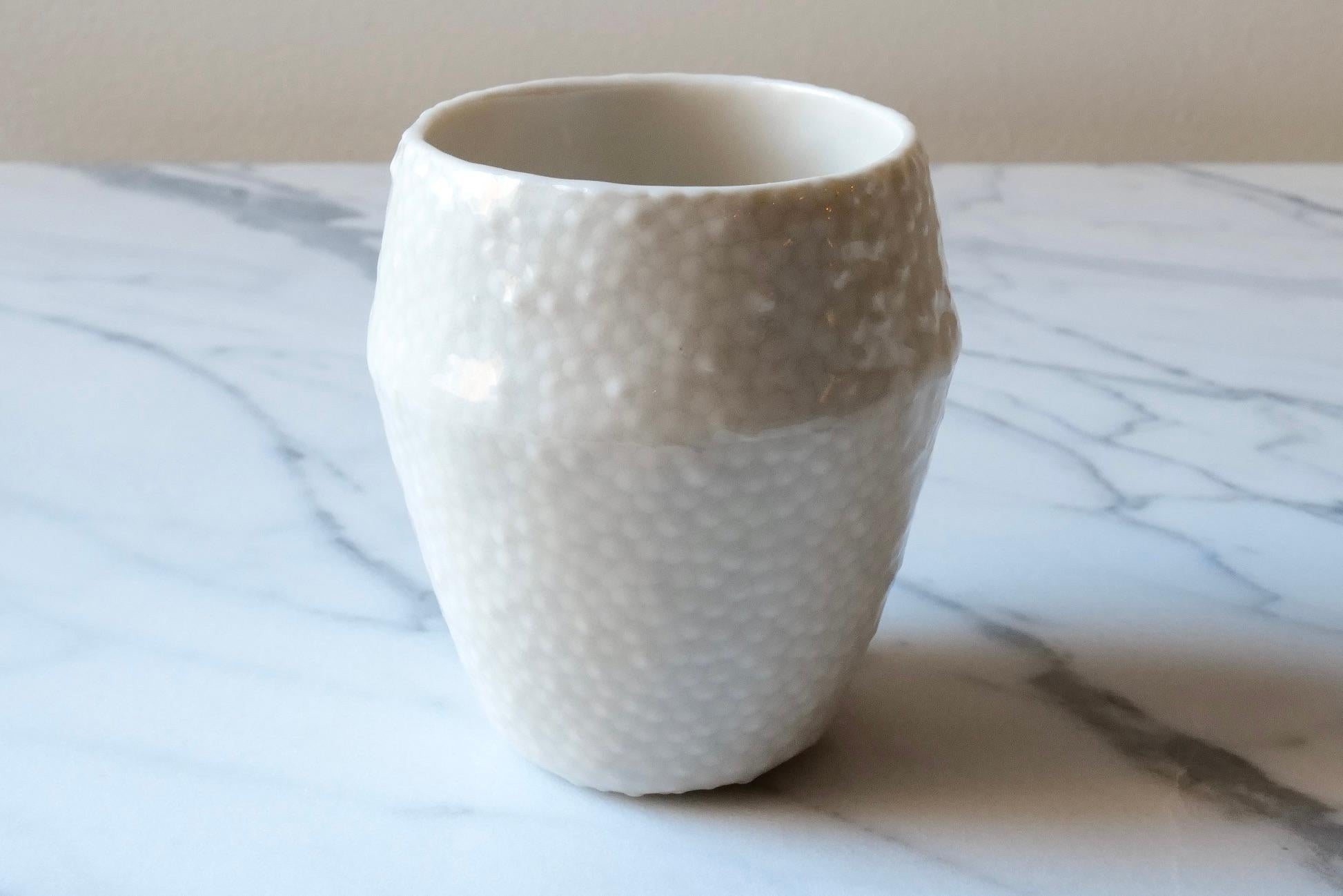 Delicate ceramic drinking cup. Hand-cast in porcelain using a mold hand-carved by the artist. The cup is coated with a thick, luminous clear glaze revealing lots of texture below a sleek glossy finish, like looking into a pond and finding frogspawn