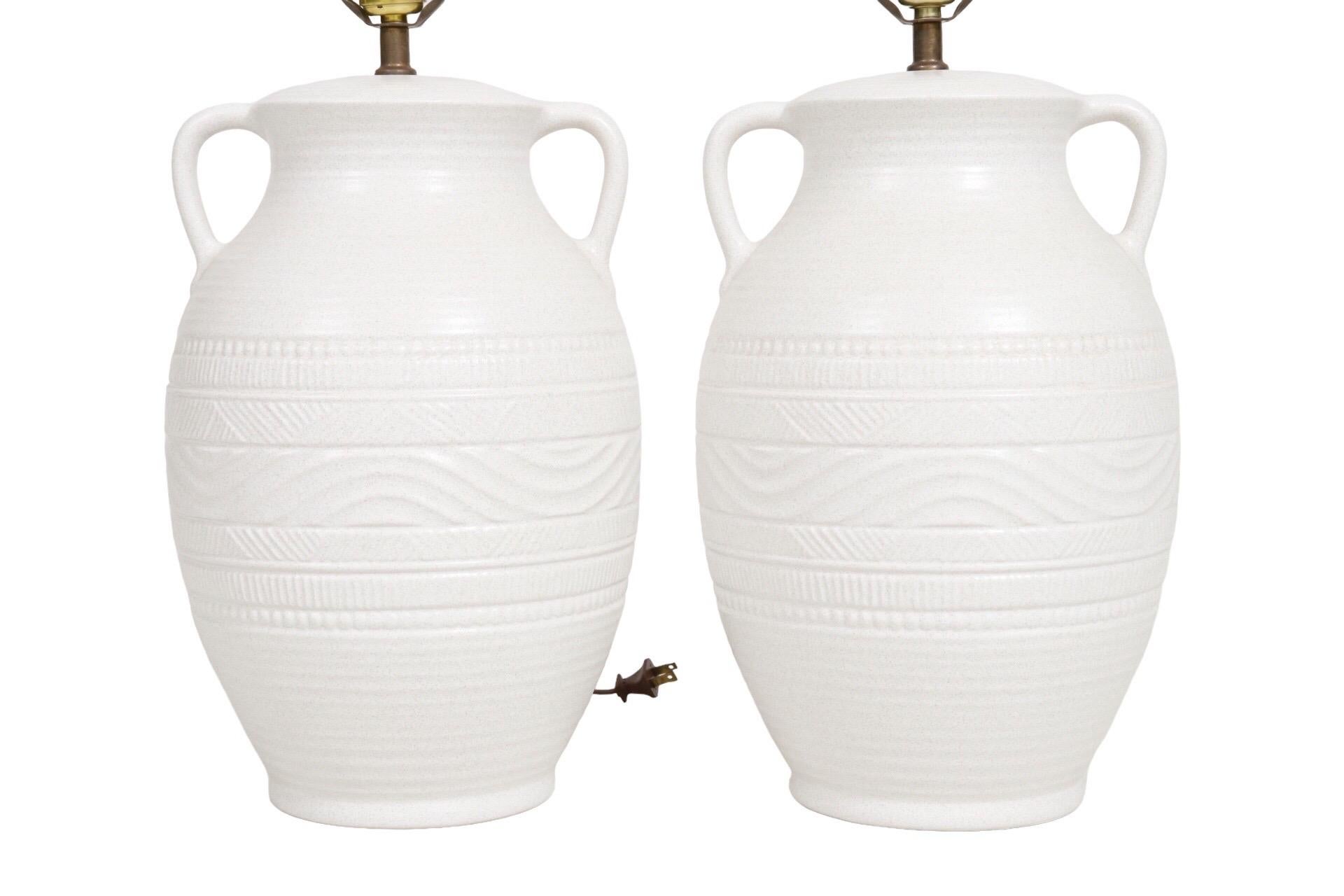 A pair of ceramic amphora shaped table lamps in white. At the neck are two small handles. Vases are pressed with undulating waves and geometric lines. Each lamp measures 9.5