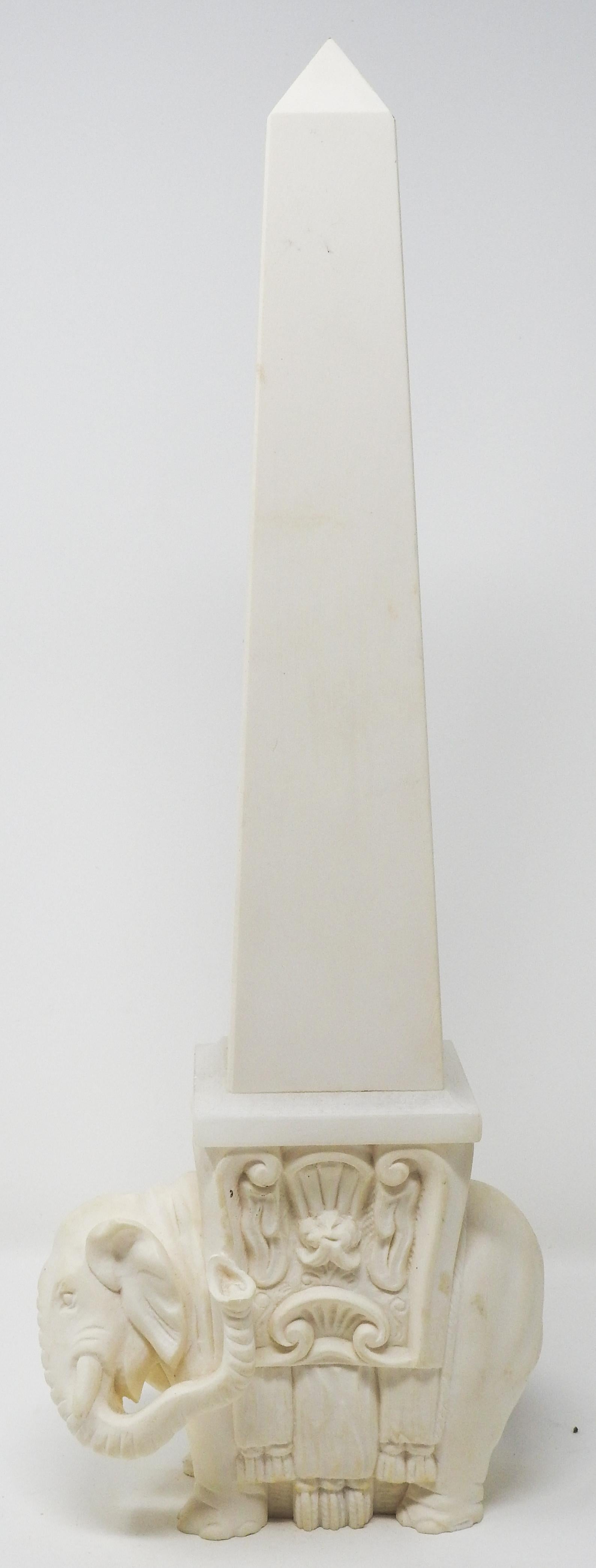 Offering this white ceramic and glass elephant obelisk. The base of this obelisk is a ceramic cast elephant that is draped with fabric and a saddle. The piece between the elephant and the top is a frosted glass piece and the top is ceramic.