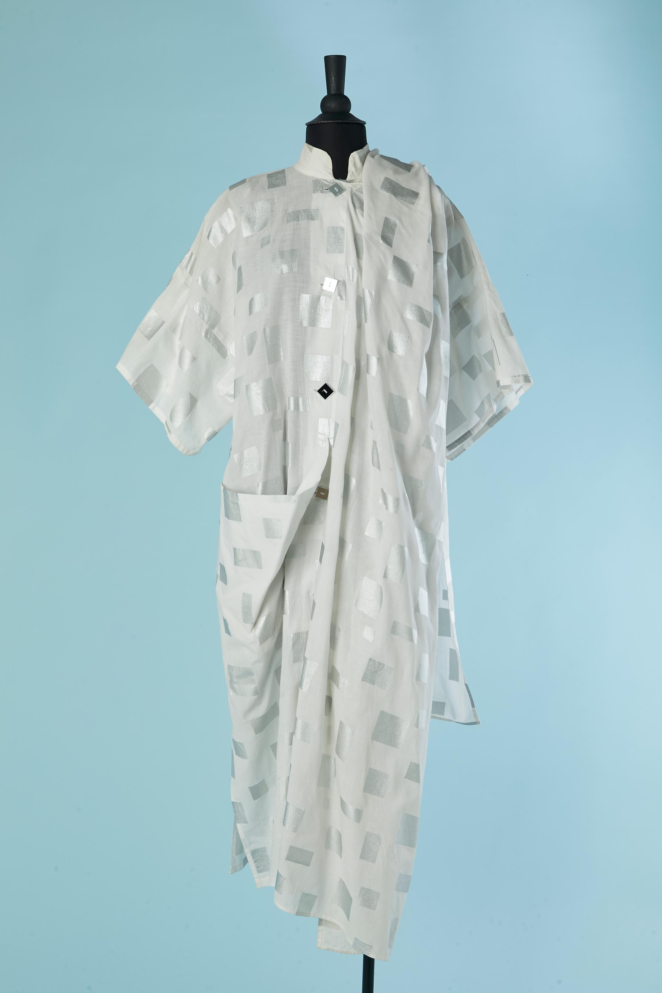 Manteau en coton blanc avec carré imprimé argenté. Bouton miroir carré et fermeture à la boutonnière sur le milieu du devant. 
Une grande poche sur le côté droit. Une sorte d'écharpe est attachée sur le côté droit et peut être enroulée autour des
