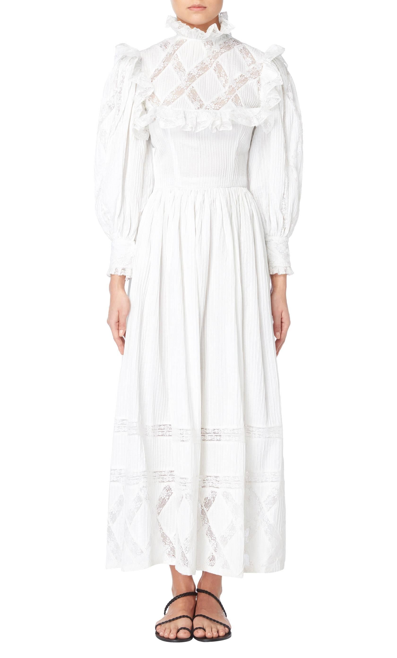 Constructed in white cotton, this high neck maxi dress features long puffed sleeves and a pie crust collar. With a fitted bodice and flared skirt, panels of sheer white lace run throughout the dress, while lace trim edges the neck and cuffs.