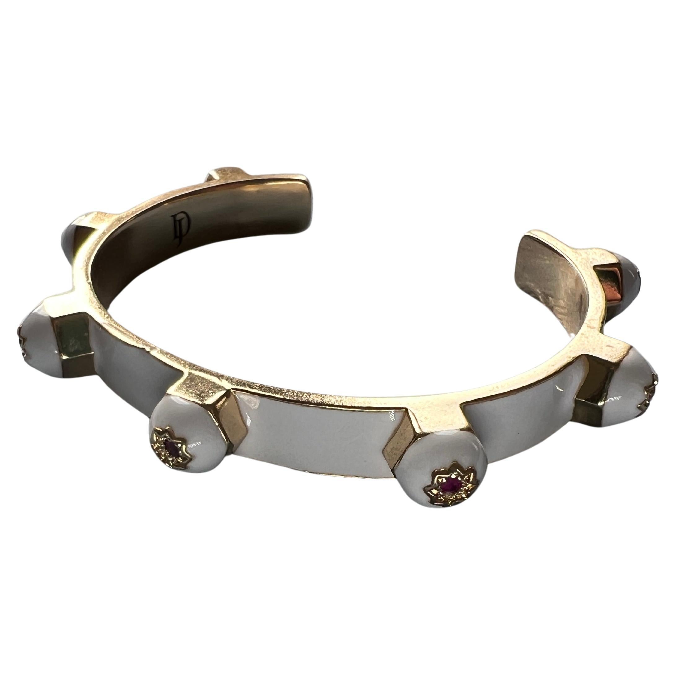 Ce bracelet manchette en émail blanc de J DAUPHIN est réalisé en laiton plaqué or 14 carats et serti de sept rubis naturels, chacun positionné dans des étoiles gravées à huit branches. La pièce allie un minimalisme épuré à un poids symbolique,