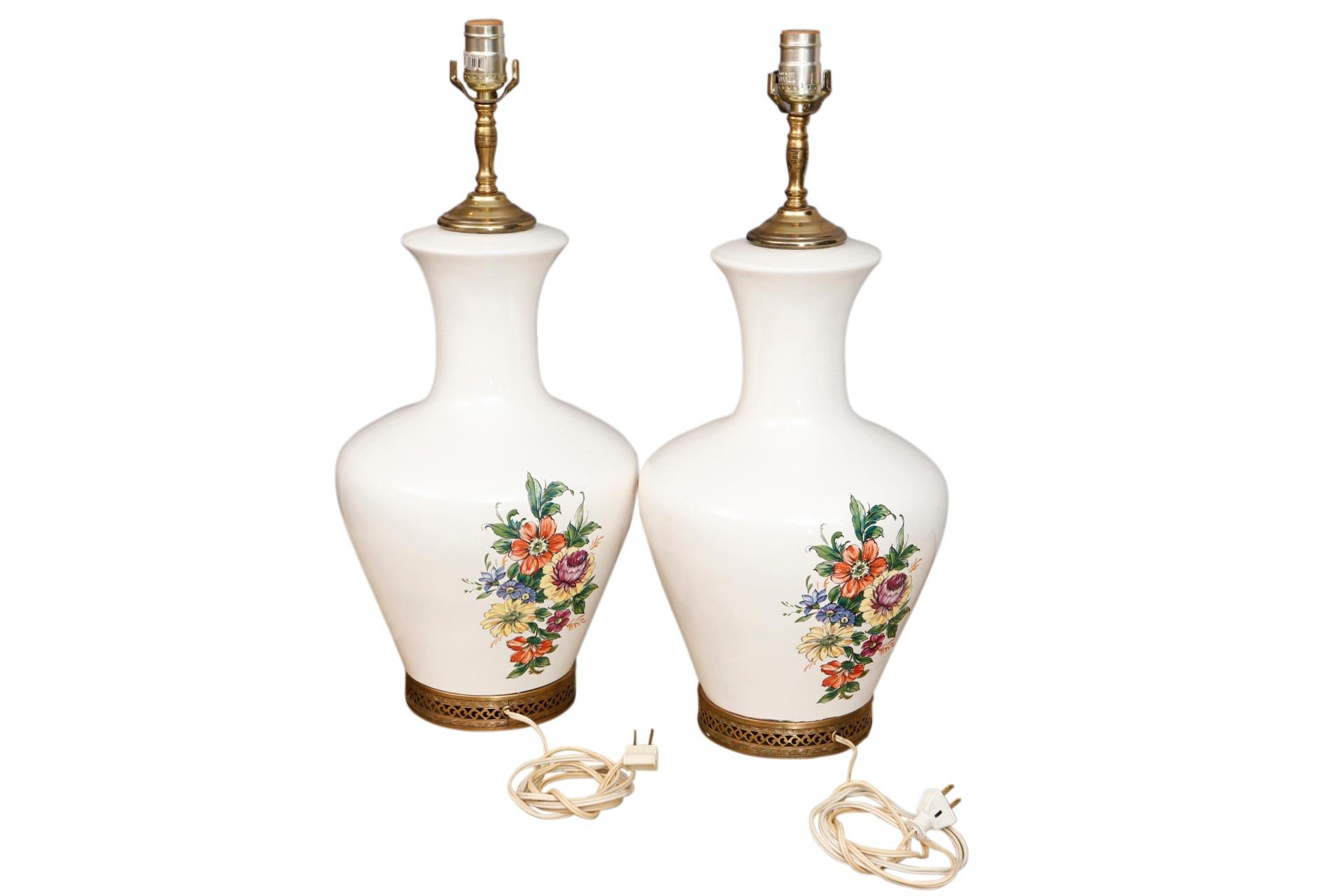 A pair of white floral table lamps. Simple ceramic white vases with slender necks and a wide vase are decorated with a delicate floral motif in blush, indigo & lilac, finished with an ornamental pierced brass base. Wired and working. Dimensions per