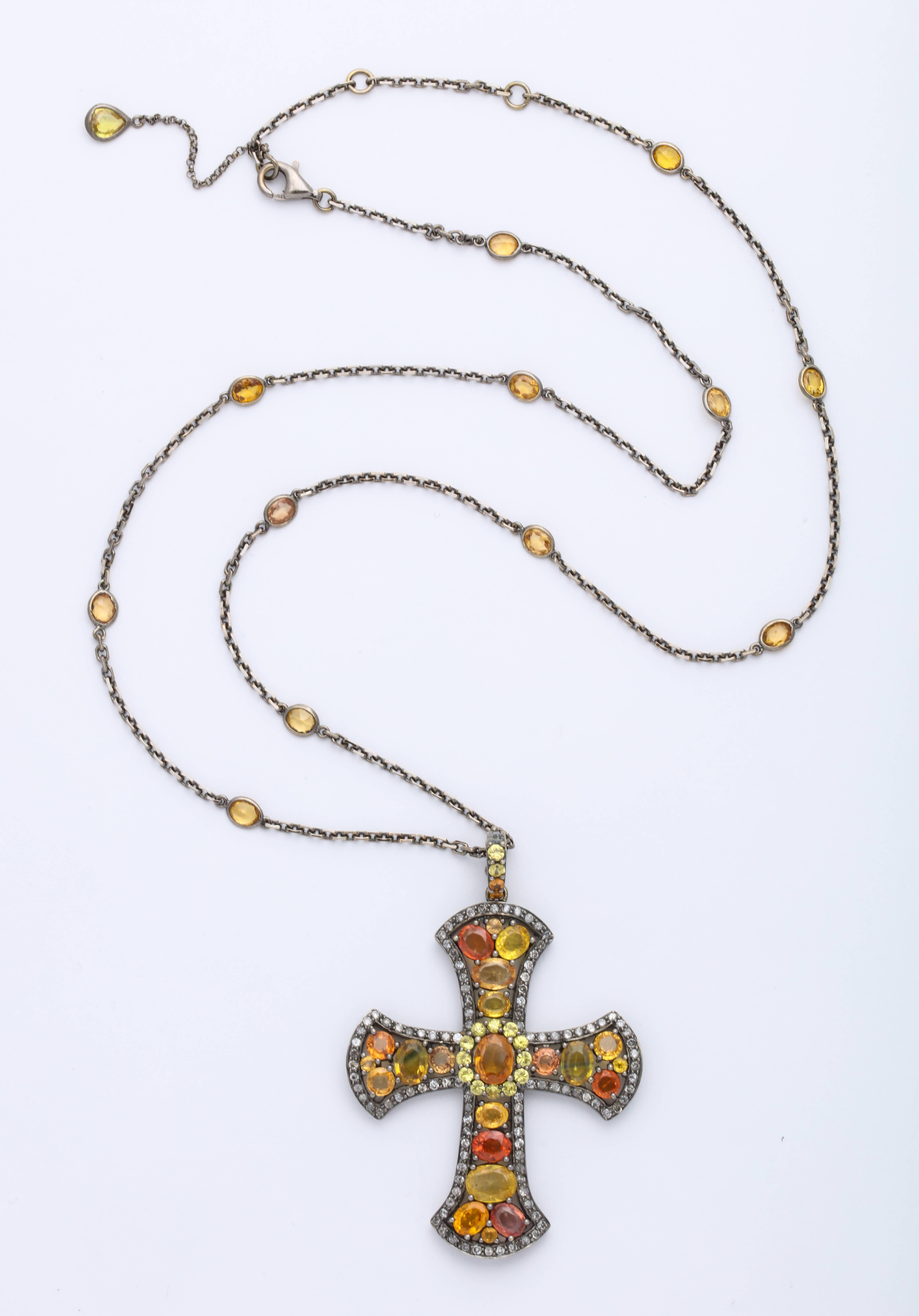 This unique 18 karat gold stylized gothic cross pendant is mounted with multi-color orange sapphires: 18.51 carats, decorated with round brilliant cut diamond trim weighing 1.65 carats, and suspended from a link chain with integrated bezel-set oval