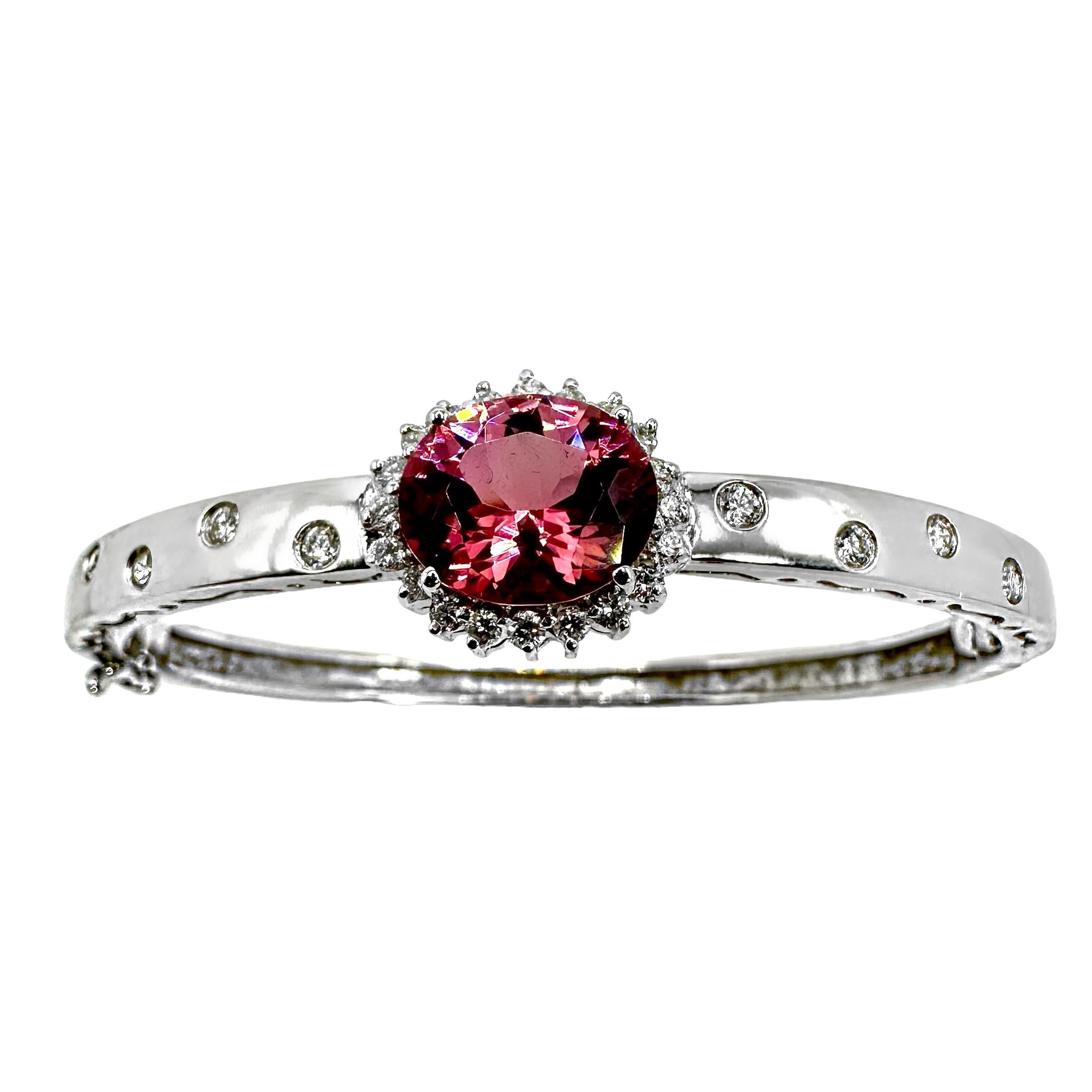 A lovely 18K white gold bangle featuring one oval shaped vibrant pink tourmaline surrounded by diamonds in a halo style setting in the center. In addition, each shoulder of this bracelet is burnished with 4 diamonds inset. The top and bottom of the