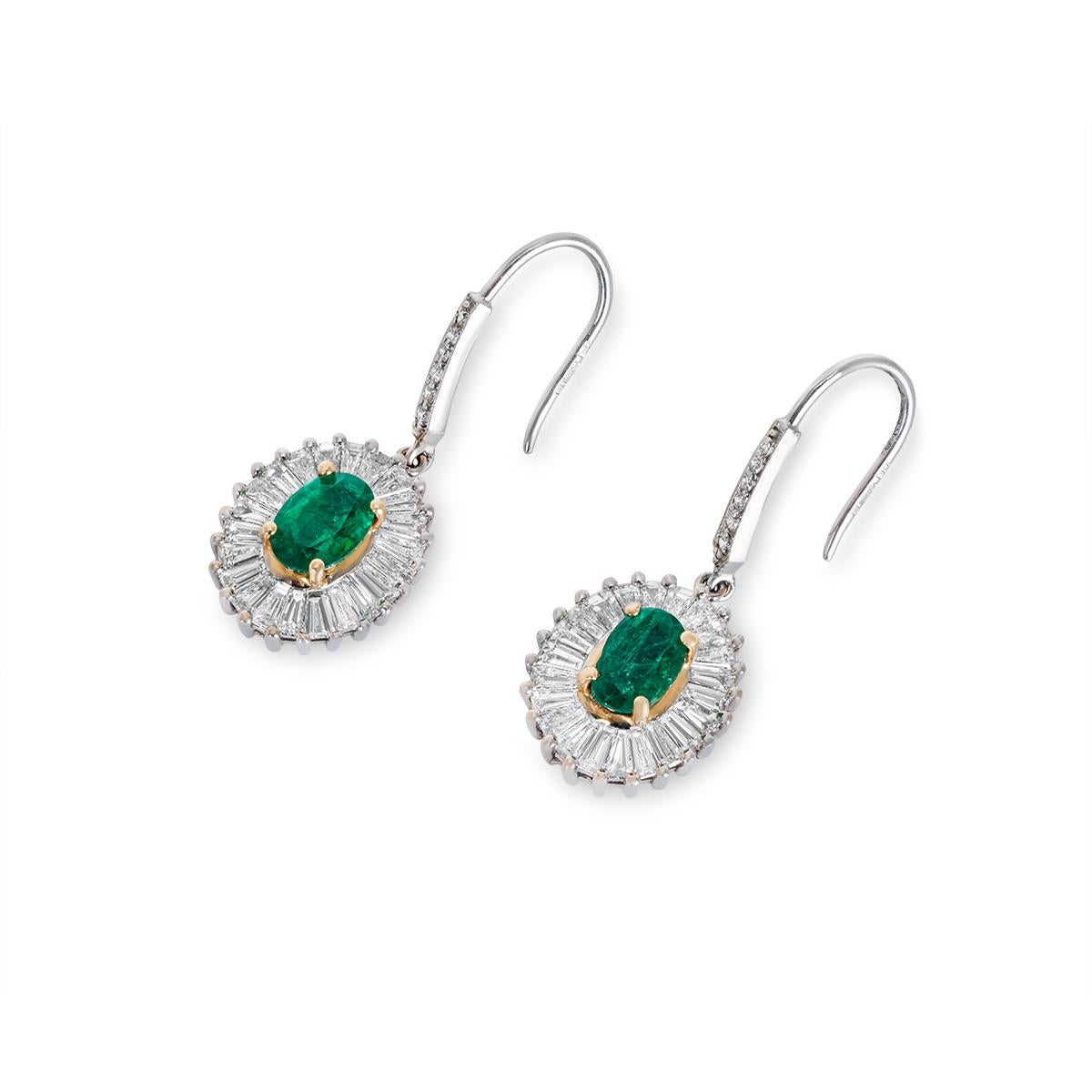 A dazzling pair of 18k white gold emerald and diamond drop earrings. The earrings highlight oval cut emeralds set to the centre with 18k yellow gold prongs with an approximate total weight of 1.63ct and displaying a vibrant green hue. Accentuating