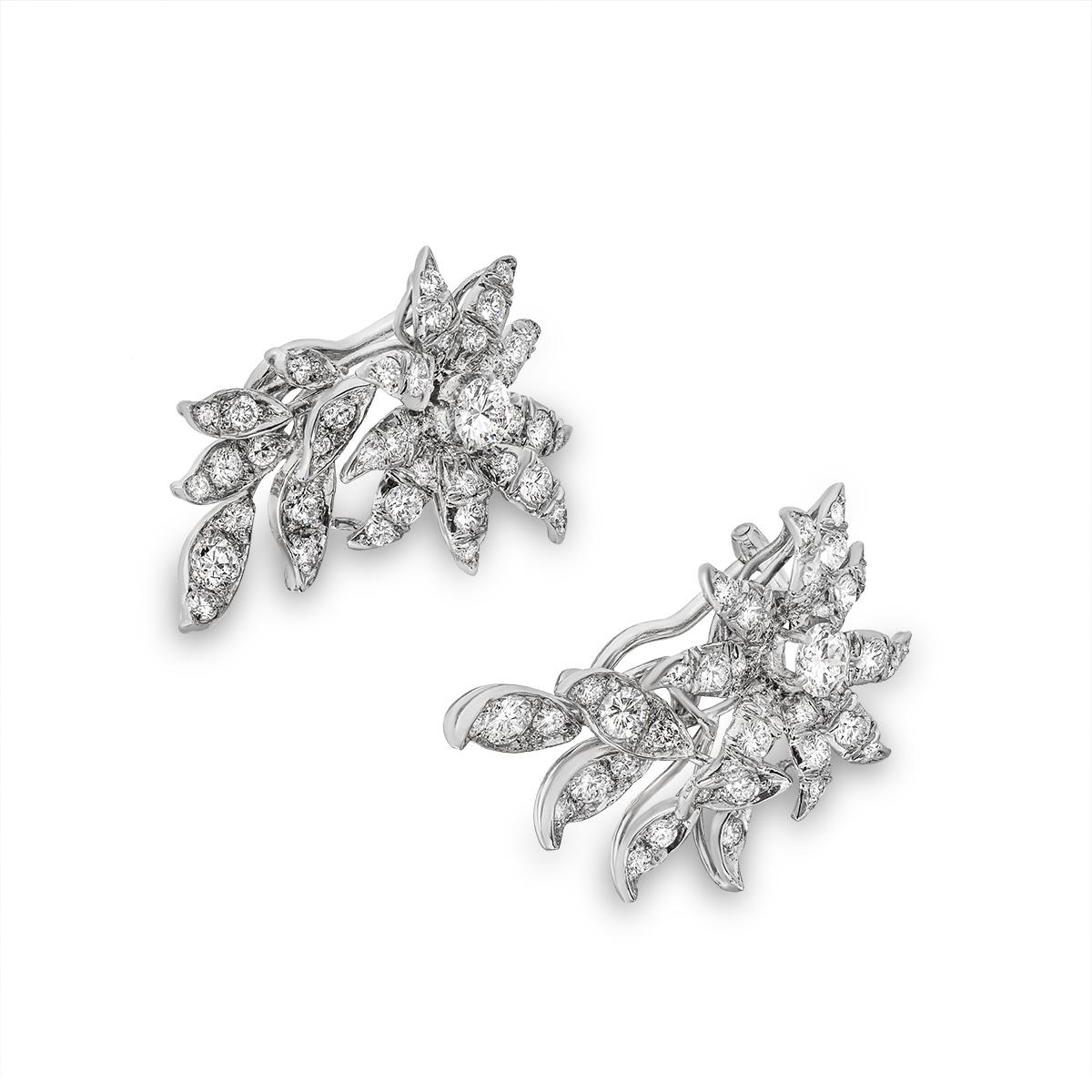 A unique pair of 18k white gold diamond clip-on earrings. The earrings feature a floral design each set to the centre with a round brilliant cut diamond weighing approximately 0.35ct, F-G colour and VS clarity. Further complementing the centre