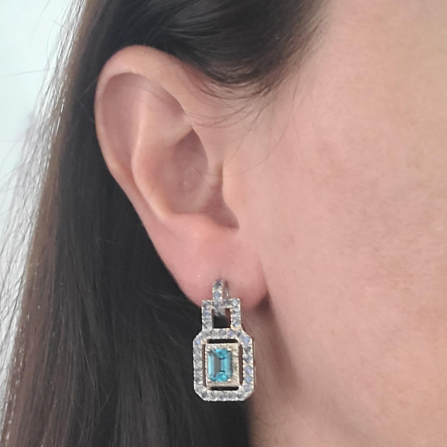 14 Karat White Gold Drop Earrings Featuring 2 Emerald Cut Blue Topaz Totaling Approximately 2.00 Carats, Accented By 36 Round Diamonds Totaling 0.18 Carats, and 72 Round Sapphires Totaling Approx. 2 Carats. 1 Inch Long. Pierced Post With Friction